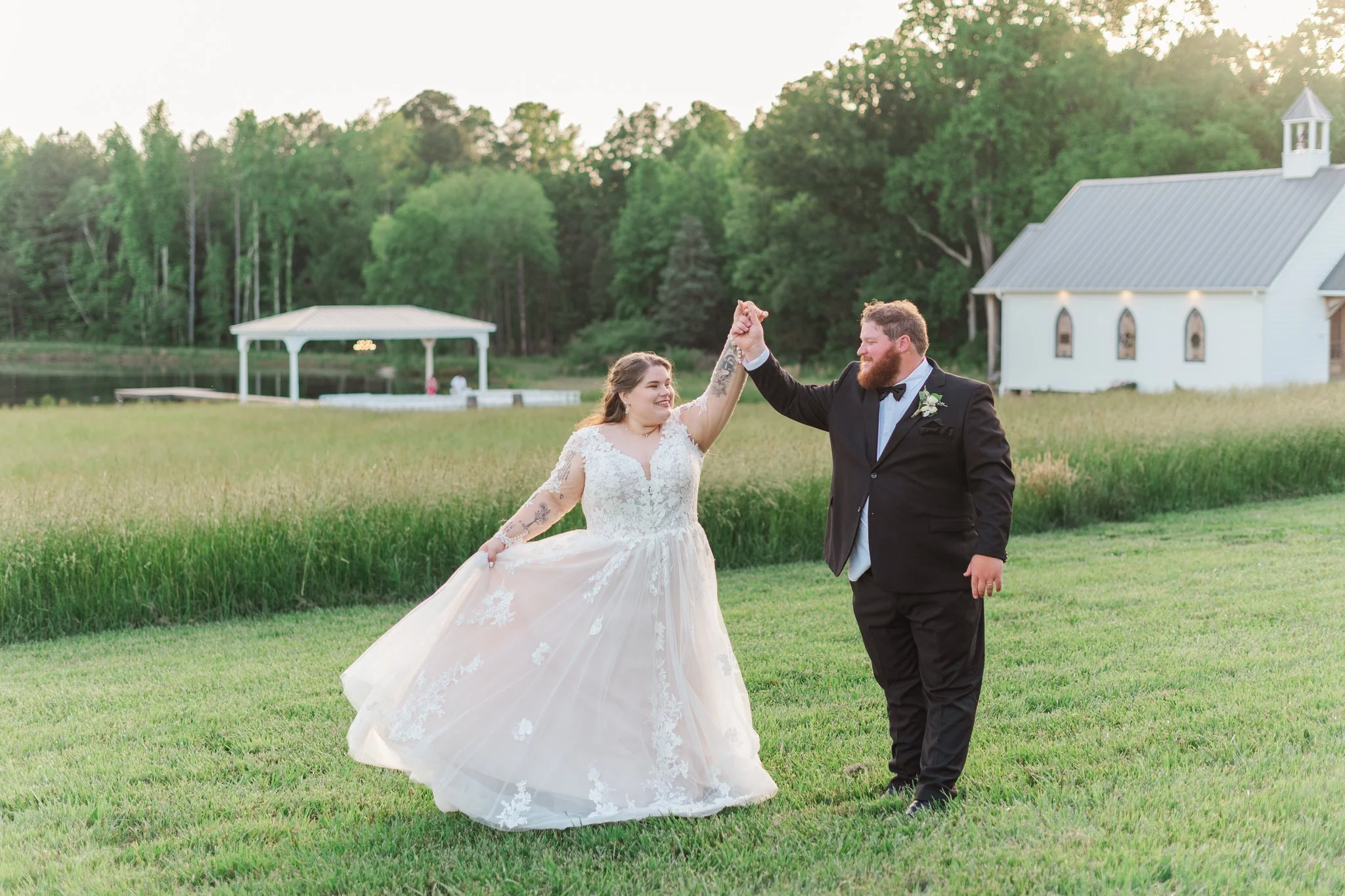 A bride and groom dancing on a grassy field during sunset, with a white church and a lake in the background.