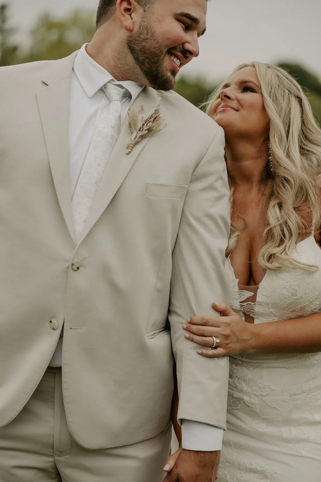 A couple dressed in wedding attire, with the man in a light beige suit and the woman in a white lace wedding dress, sharing a joyful moment outdoors.