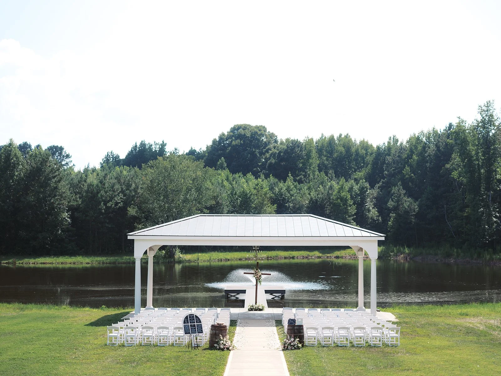 Outdoor wedding ceremony setup with white chairs, a small white pavilion, a cross on a stand, and a lake with fountain, surrounded by trees.