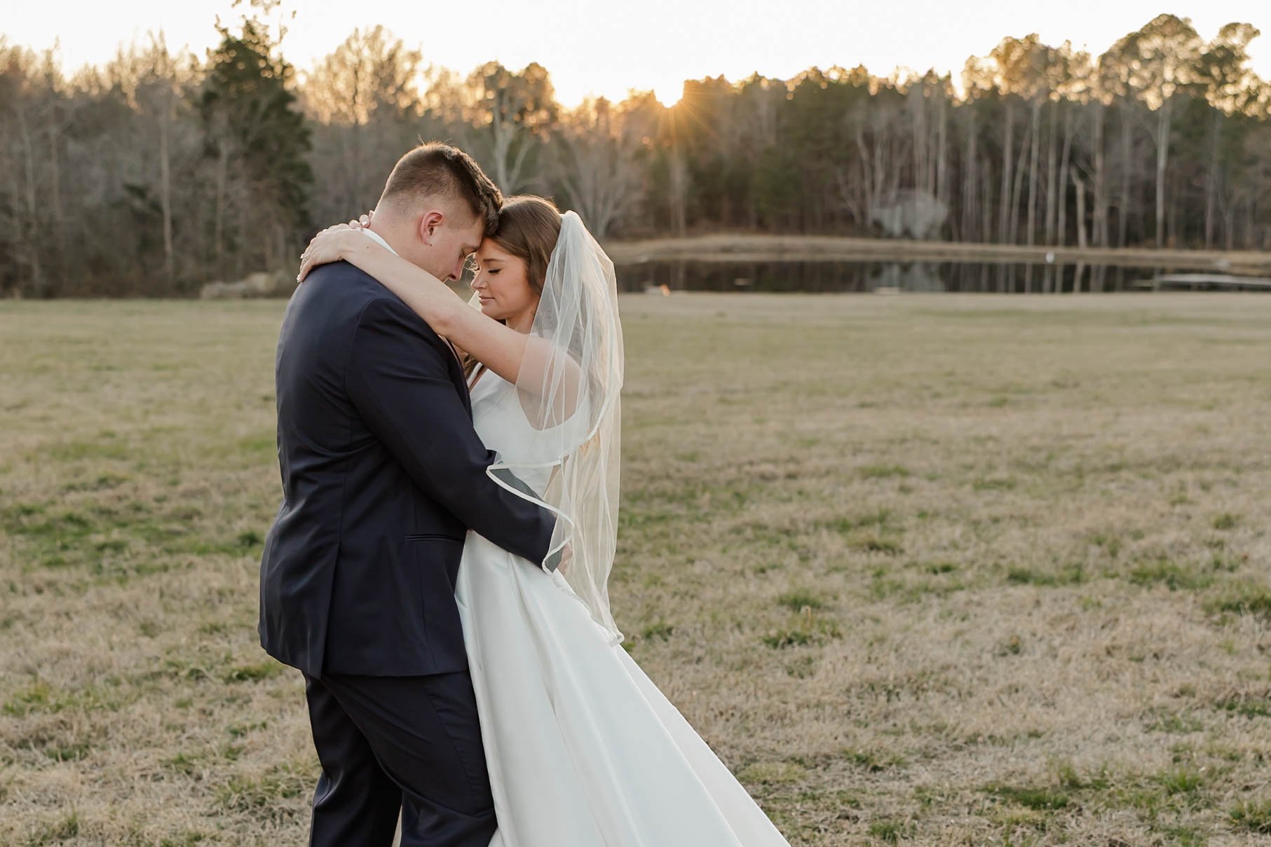 A bride and groom embrace in a field at sunset, with trees and a pond in the background.