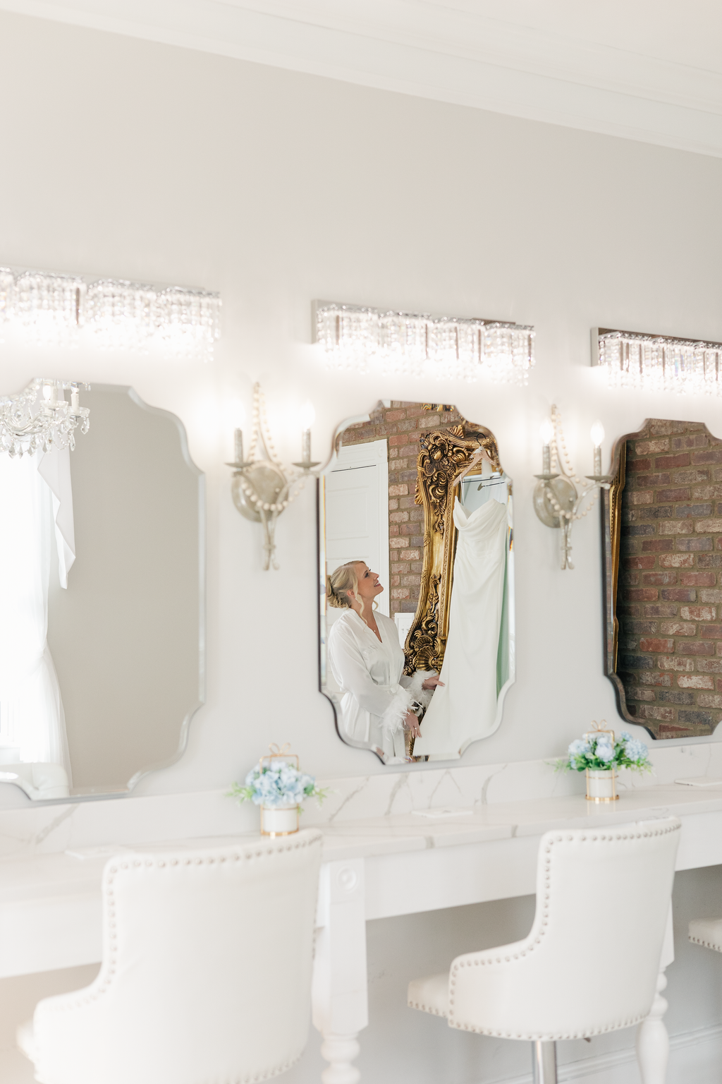 A woman in a white robe looking into a mirror with a gold ornate frame, reflected in a series of three mirrors above a white marble vanity, decorated with flower arrangements, wall sconces, and chandeliers in a bright room with white walls and a bric