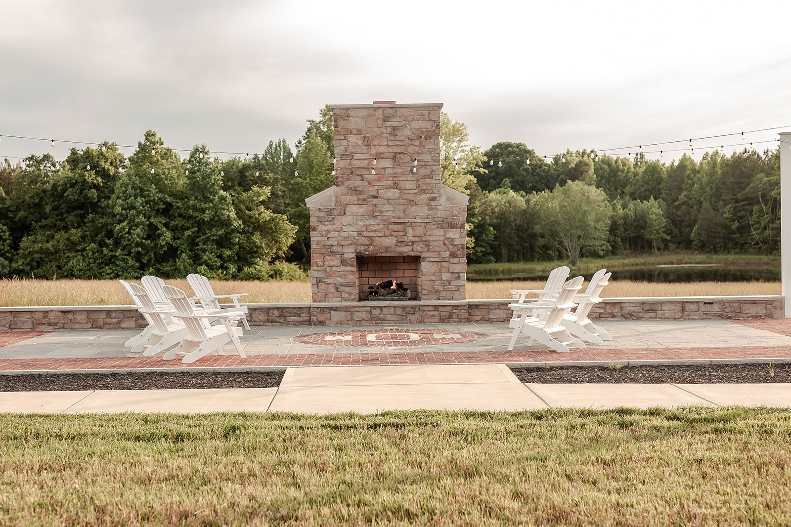 Outdoor patio with a central brick fireplace, surrounded by six white Adirondack chairs, overlooking a grassy field and trees in the background.