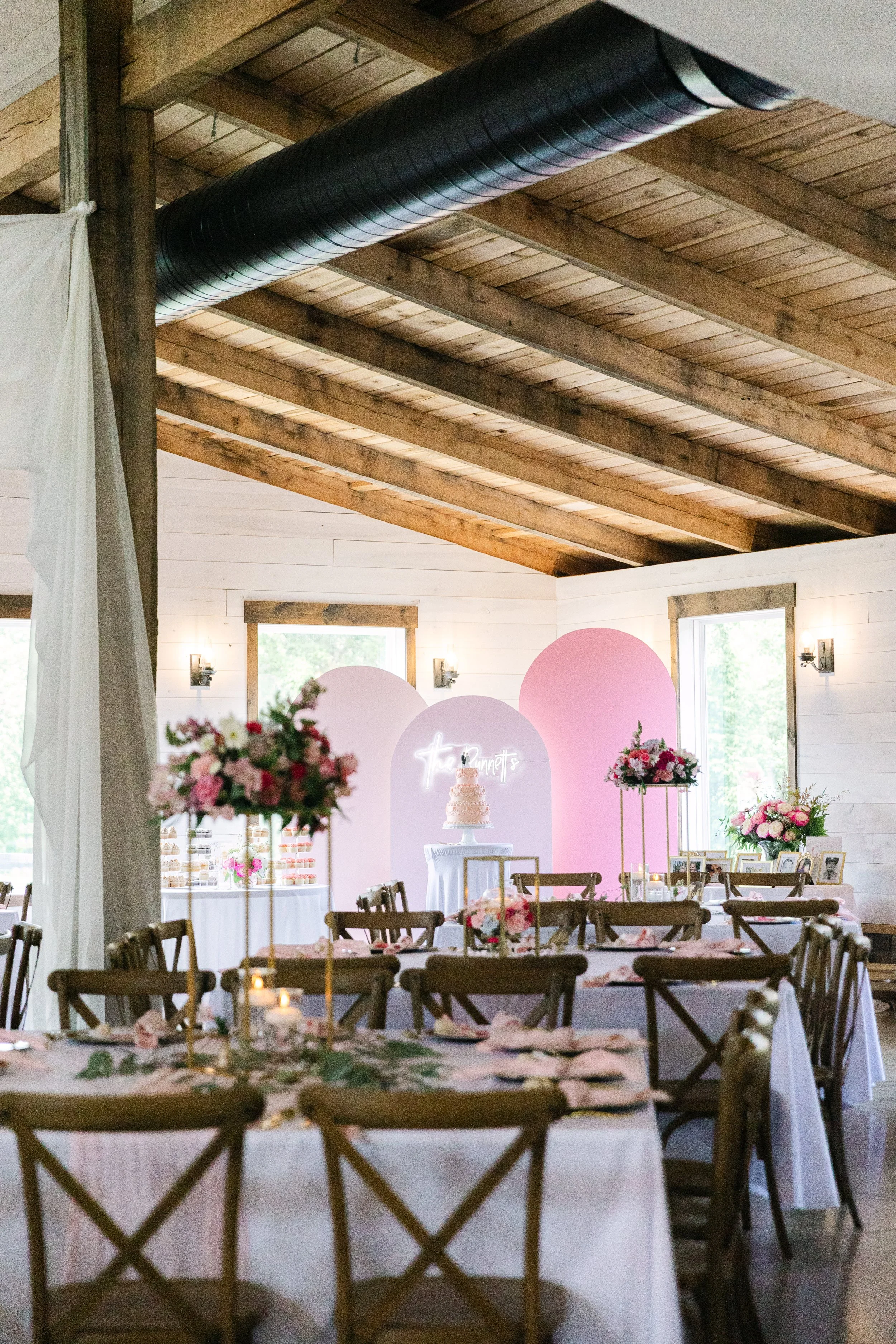 A decorated event space with wooden ceiling beams, tables with white tablecloths, pink floral centerpieces, and a dessert table with a pink and white cake, pink flowers, and framed photos. Pink and white backdrop with neon sign reading 'favourites' i