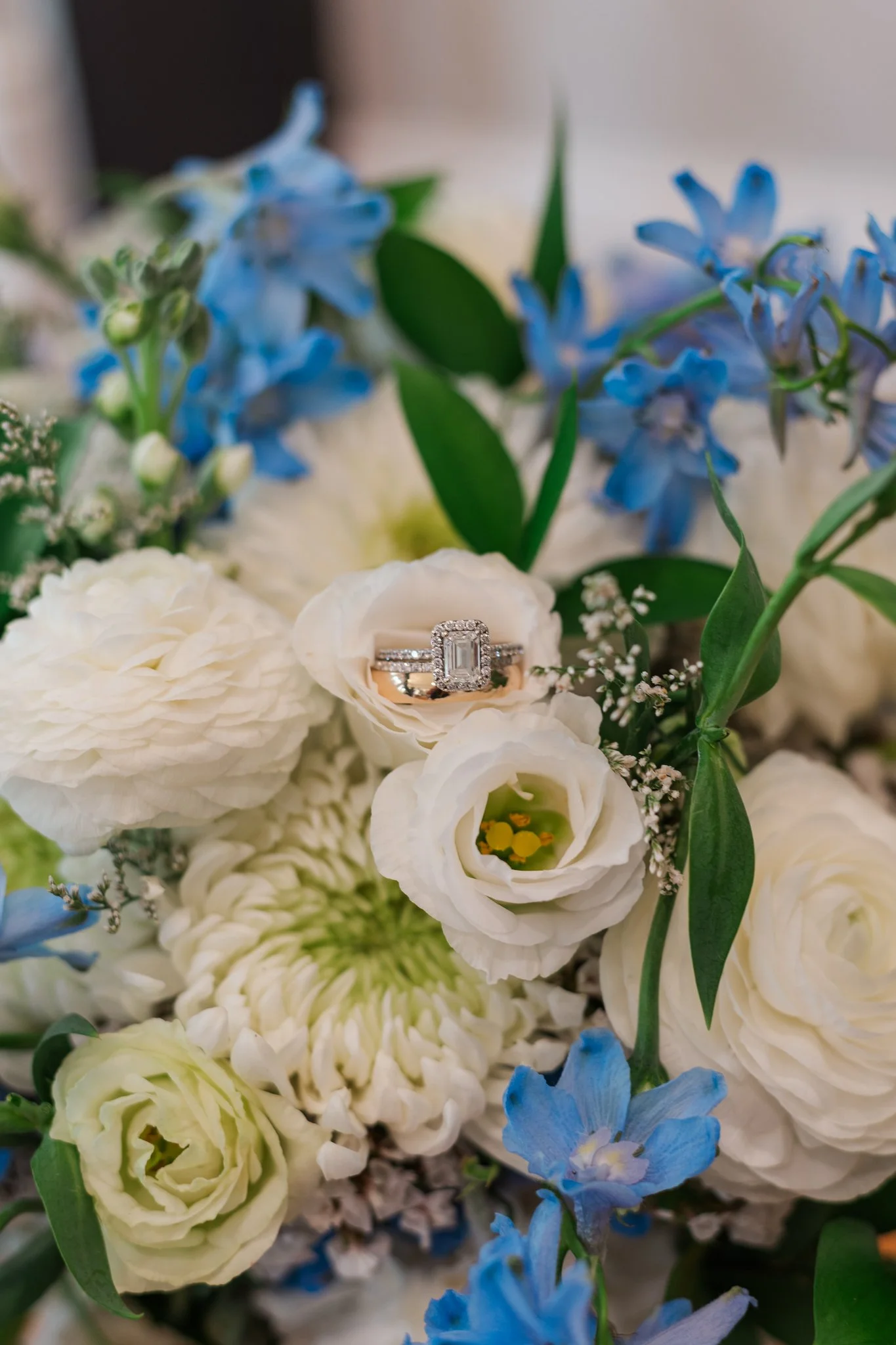A diamond engagement ring with an emerald cut center stone surrounded by smaller diamonds, resting in a bouquet of white and blue flowers.