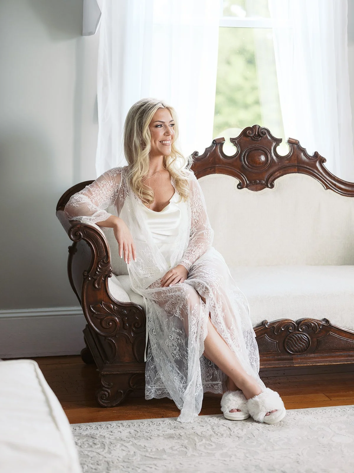 A woman with long blonde hair sitting on a vintage wooden sofa, wearing a lace robe and fluffy slippers, smiling and looking to the side near a large window with white curtains.