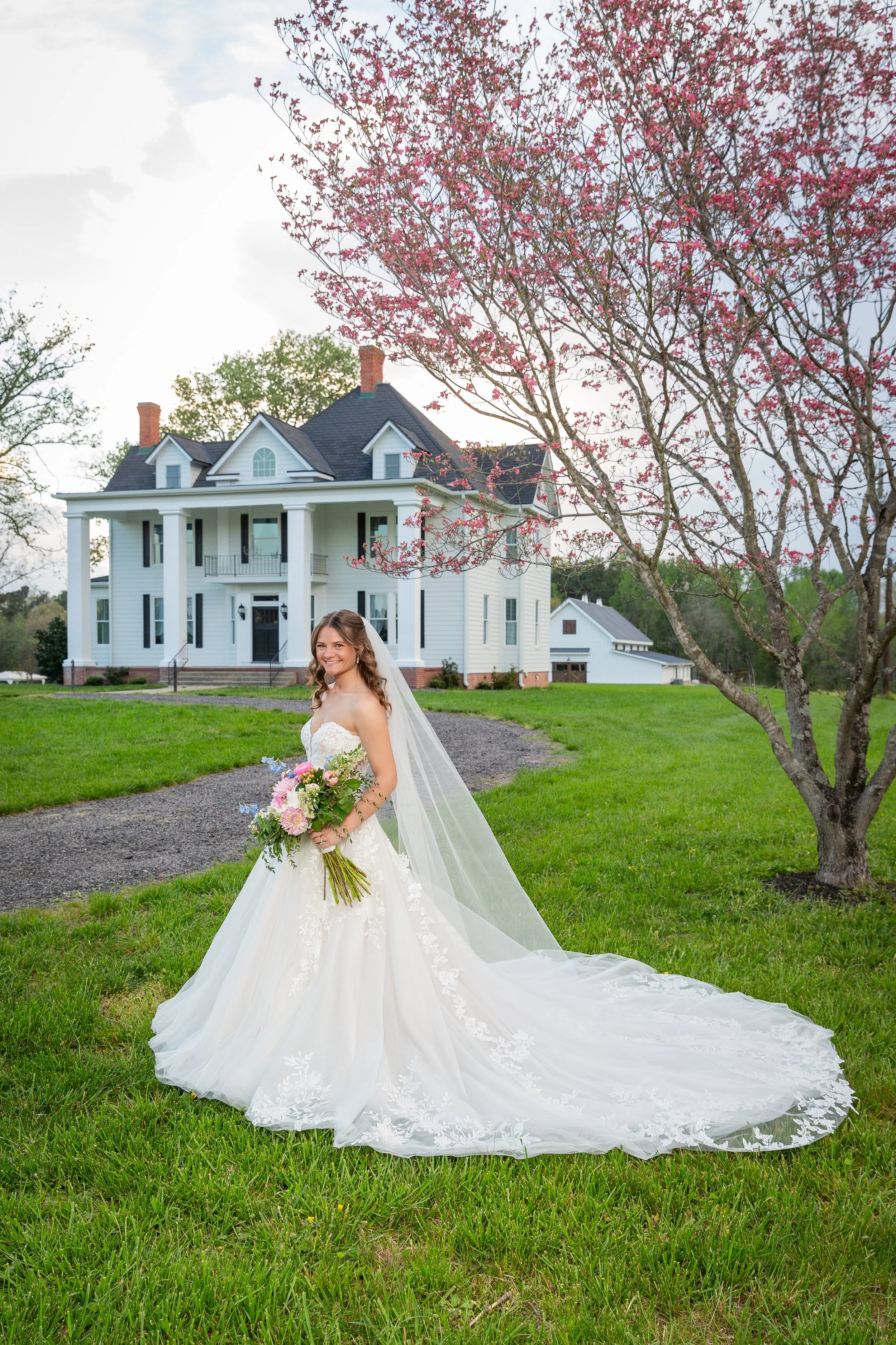 Bride in a white wedding gown holding a bouquet of pink and purple flowers, standing on green grass near a blooming pink tree, in front of a large white house with a dark roof and brick steps.