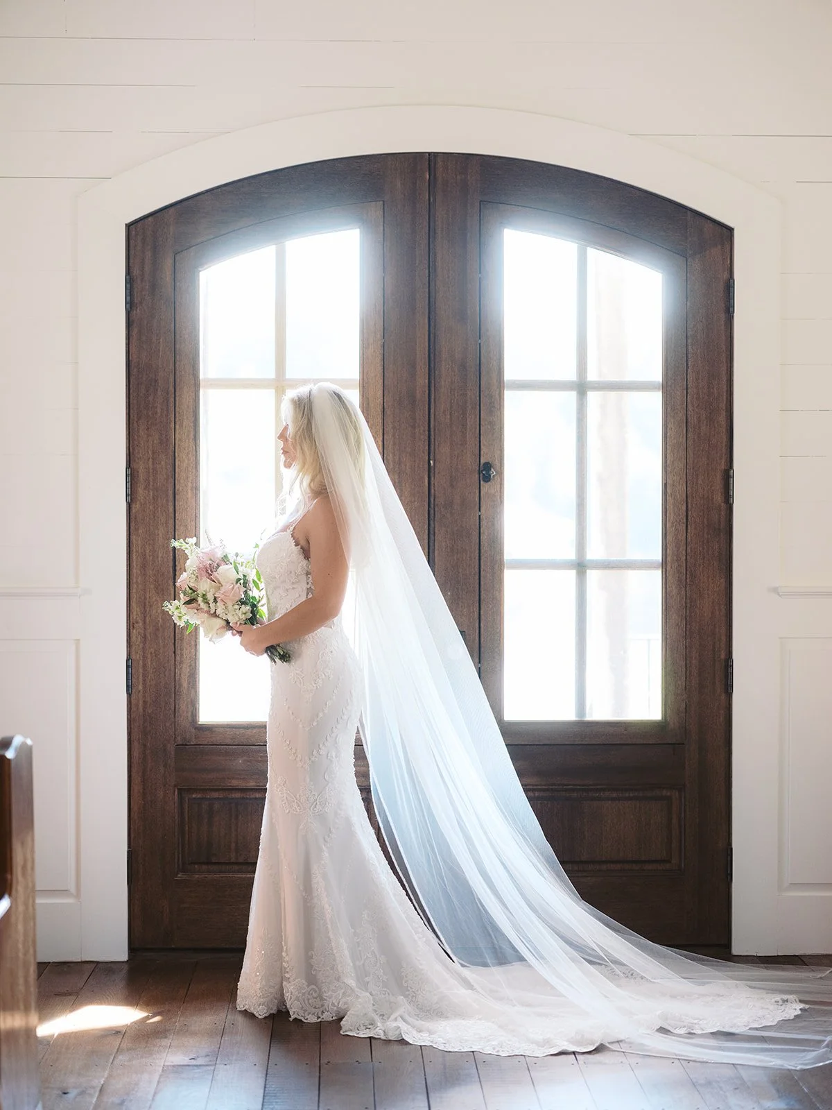 A bride in a white lace wedding dress with a long veil holding a bouquet of pink and white flowers standing in front of a large wooden arched door with windows, inside a room with light-colored walls and wooden floors.