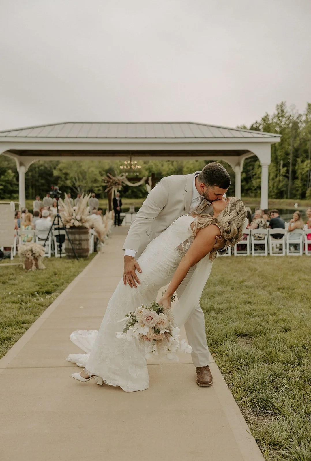 A bride and groom kissing during their outdoor wedding ceremony on a grassy area with a gazebo and guests seated on white chairs in the background.