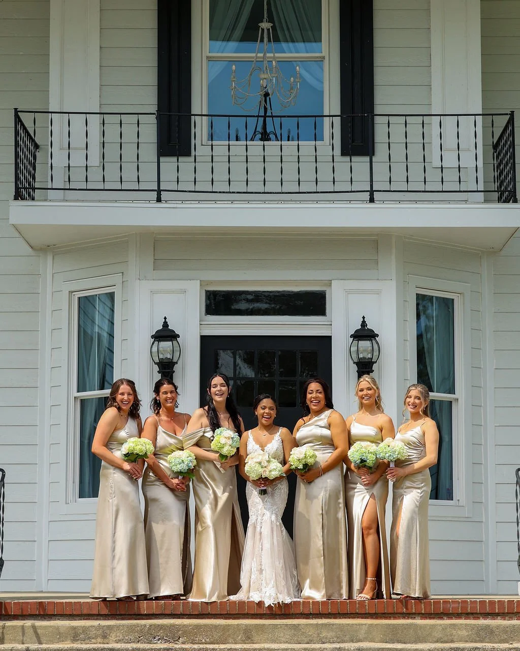 A bride and six bridesmaids standing in front of a house, all holding bouquets of white and green flowers, smiling and dressed in matching champagne-colored gowns.