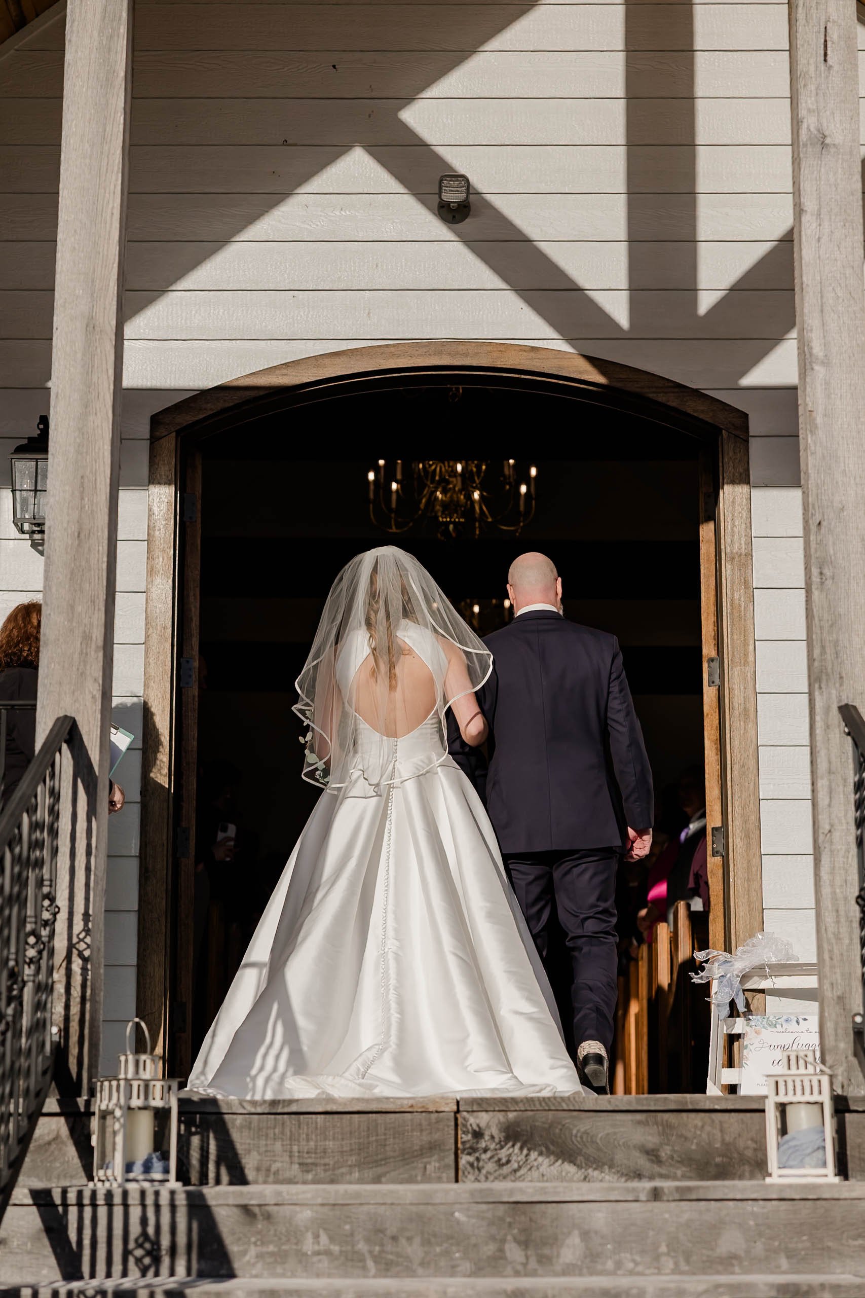 Bride and groom walking out of a church, seen from behind, with lanterns on the steps and a chandelier visible inside.