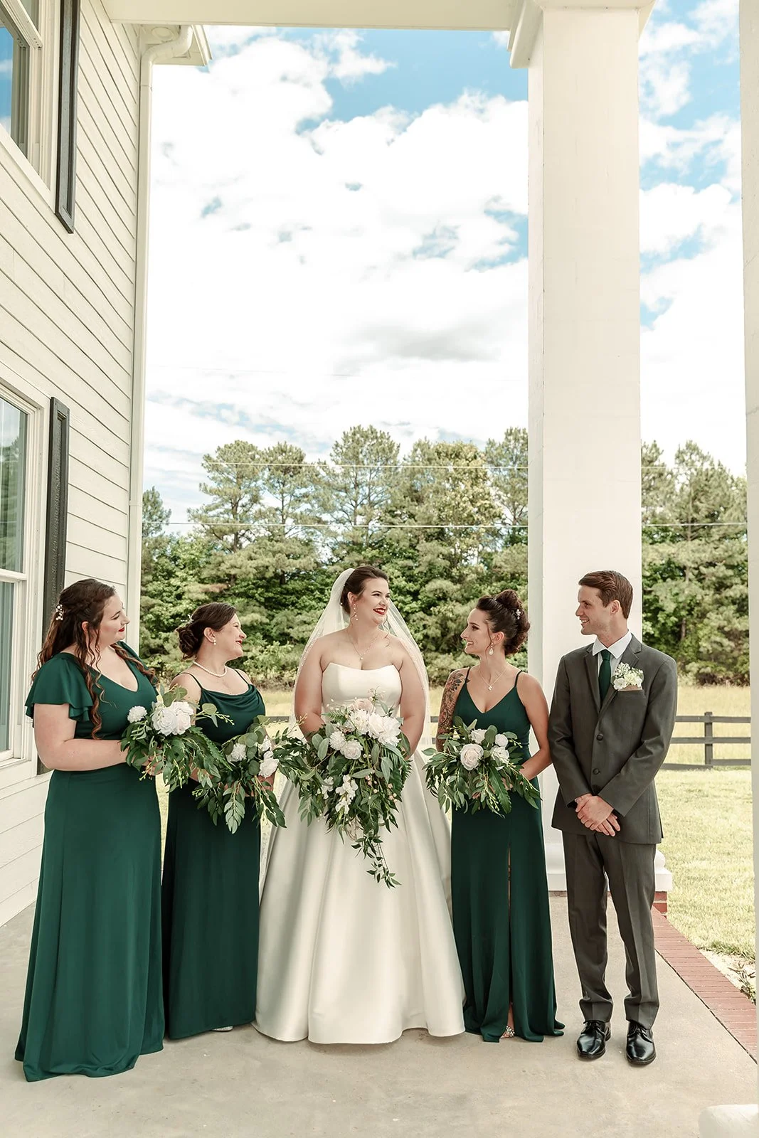 A wedding party consisting of five people, including a bride in a white gown holding a bouquet, three bridesmaids in green dresses holding bouquets, and a groom in a gray suit with a boutonniere, standing outdoors on a porch with a white house and tr