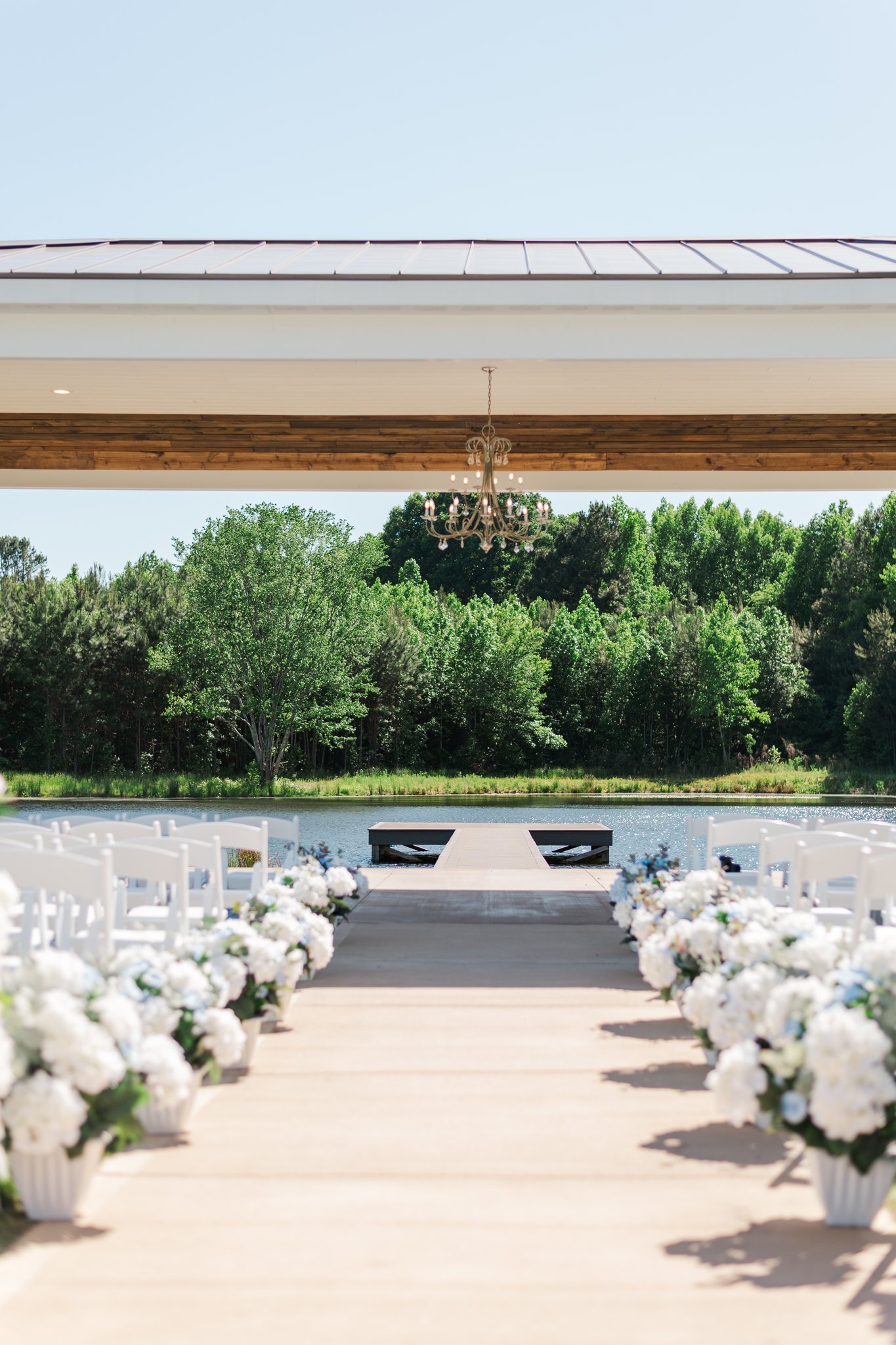 An outdoor wedding setup by a water body with white chairs and floral arrangements, overlooking a wooded area under a roofed structure with a chandelier.