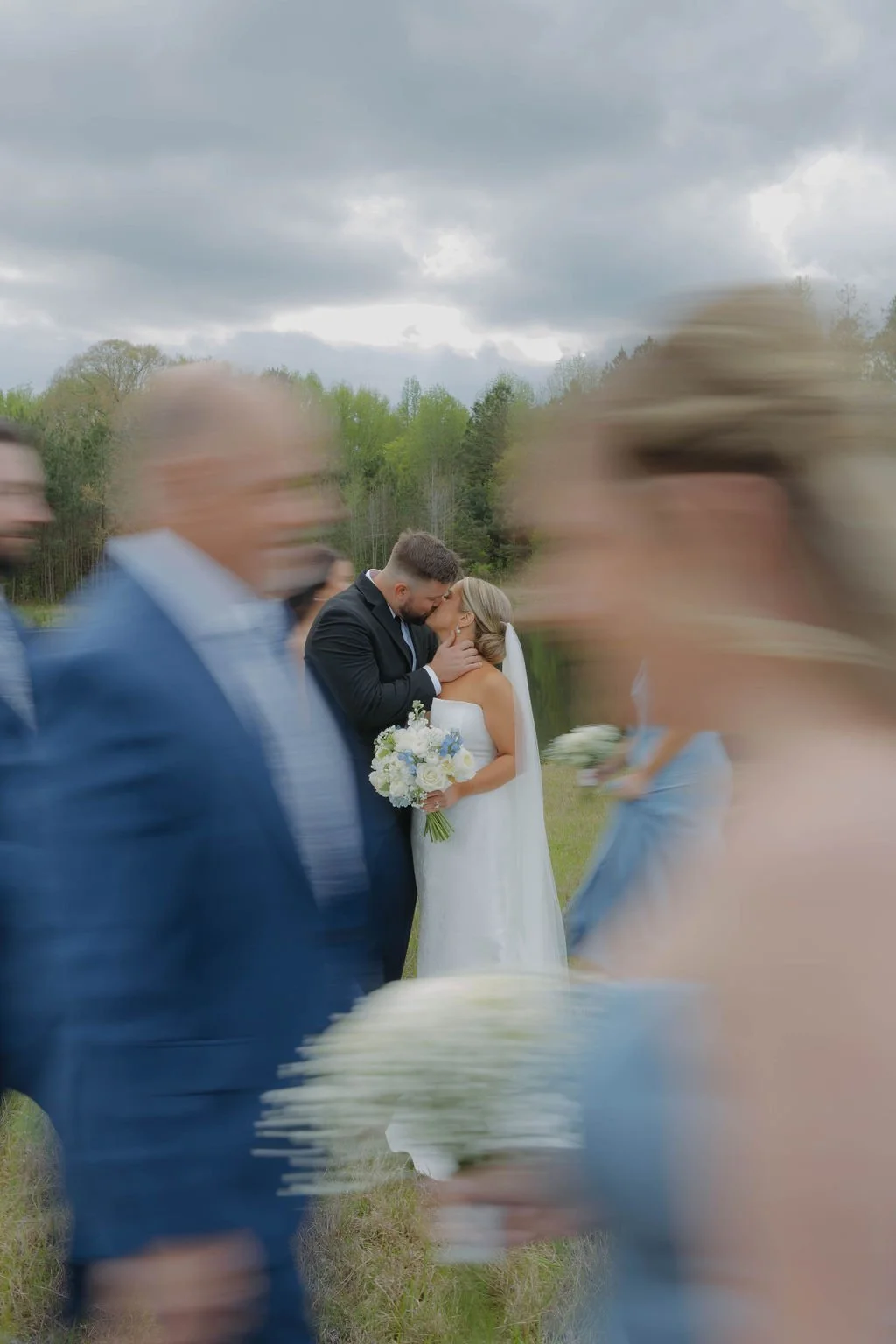 A wedding ceremony outdoors with the bride and groom sharing a kiss, holding a bouquet of flowers, while blurred guests stand in the foreground against a cloudy sky and green trees in the background.