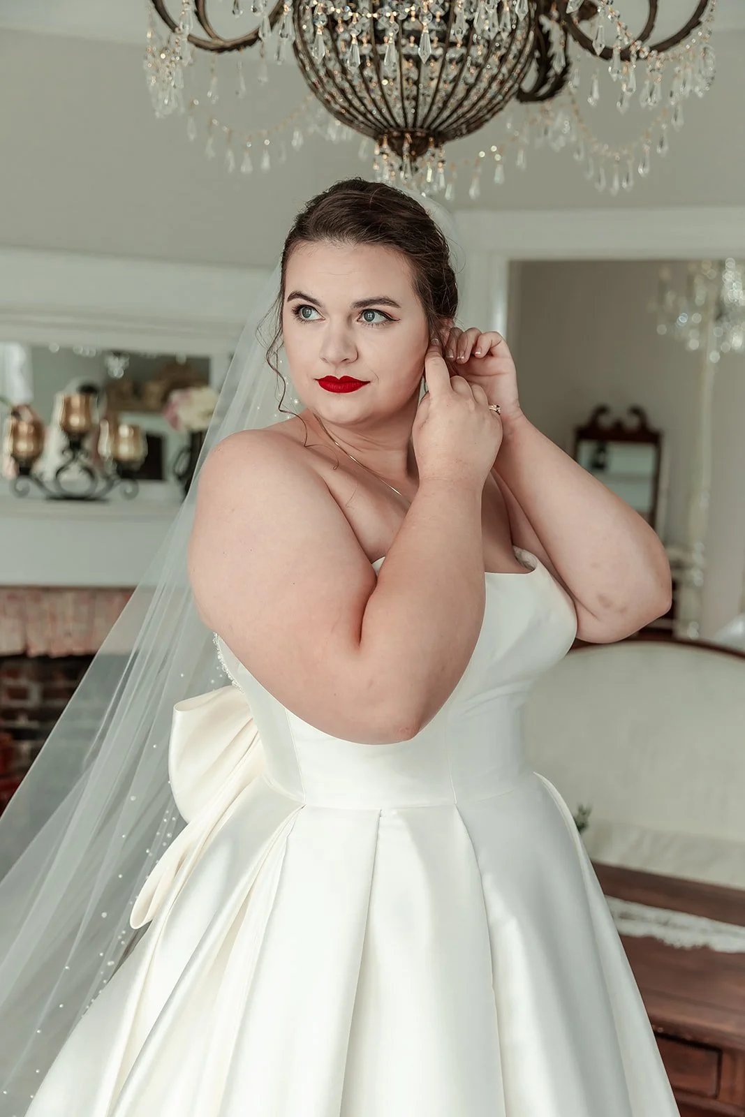 A bride in a white wedding dress adjusting her earring in a decorated room with a chandelier overhead.