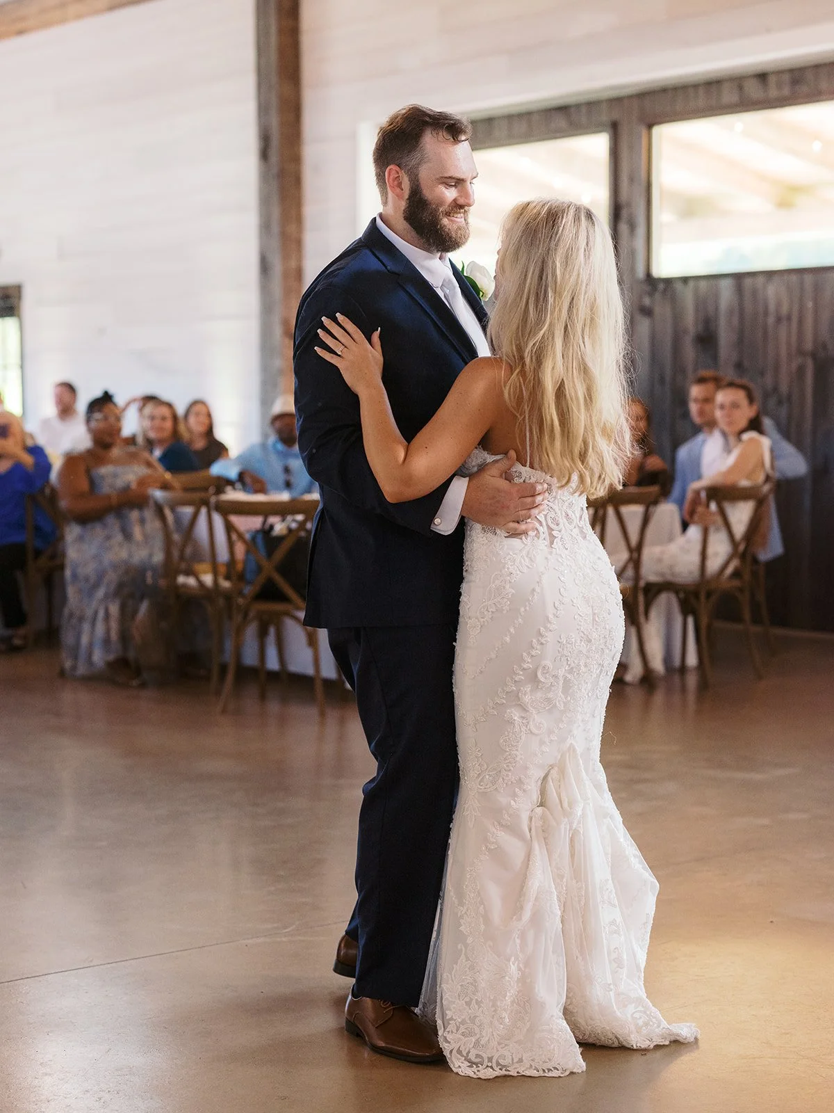 A bride and groom are dancing together at their wedding reception with guests seated at tables in the background.