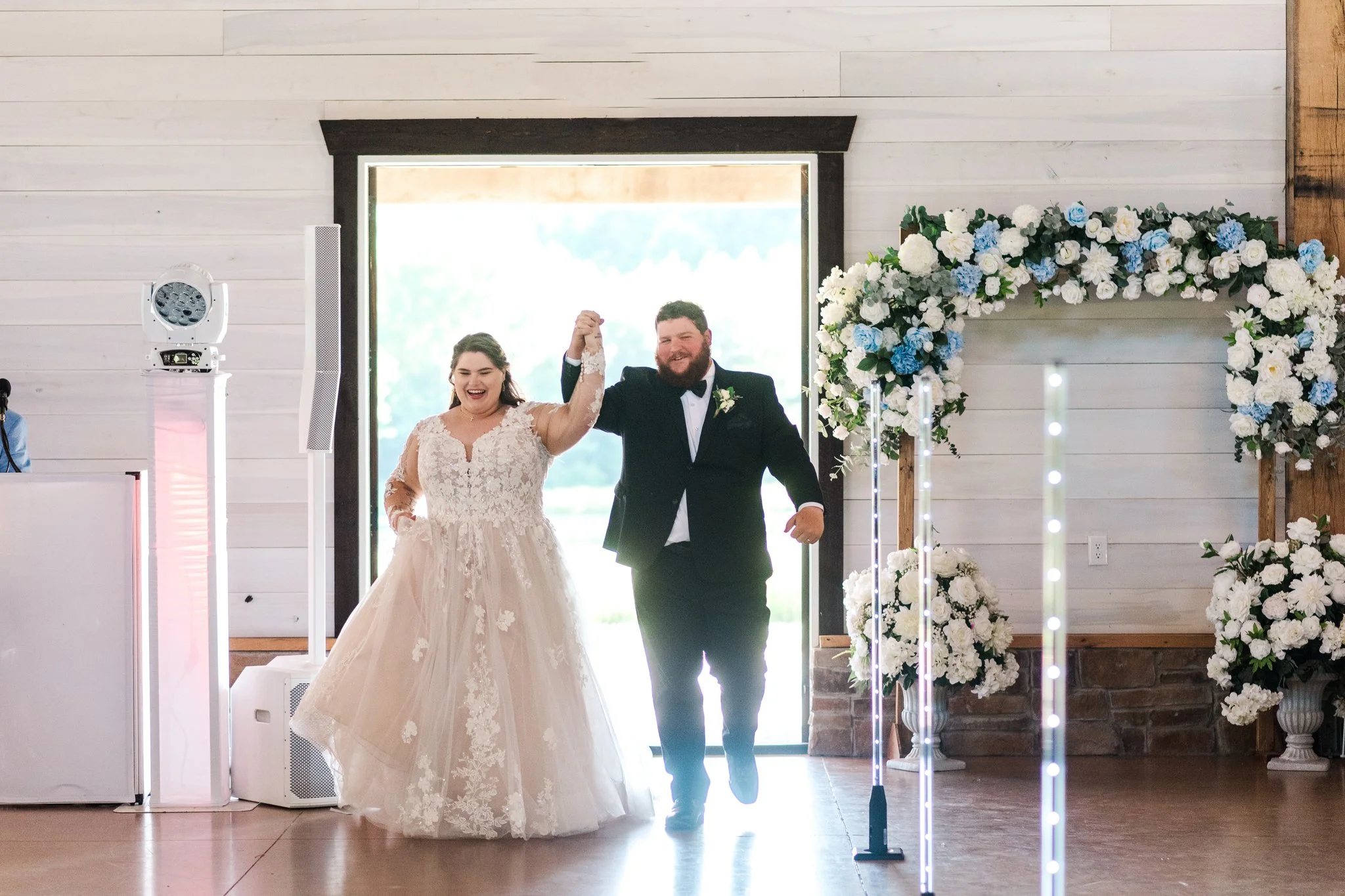 A bride and groom entering a wedding venue, holding hands and smiling, with floral decorations and a bright background.