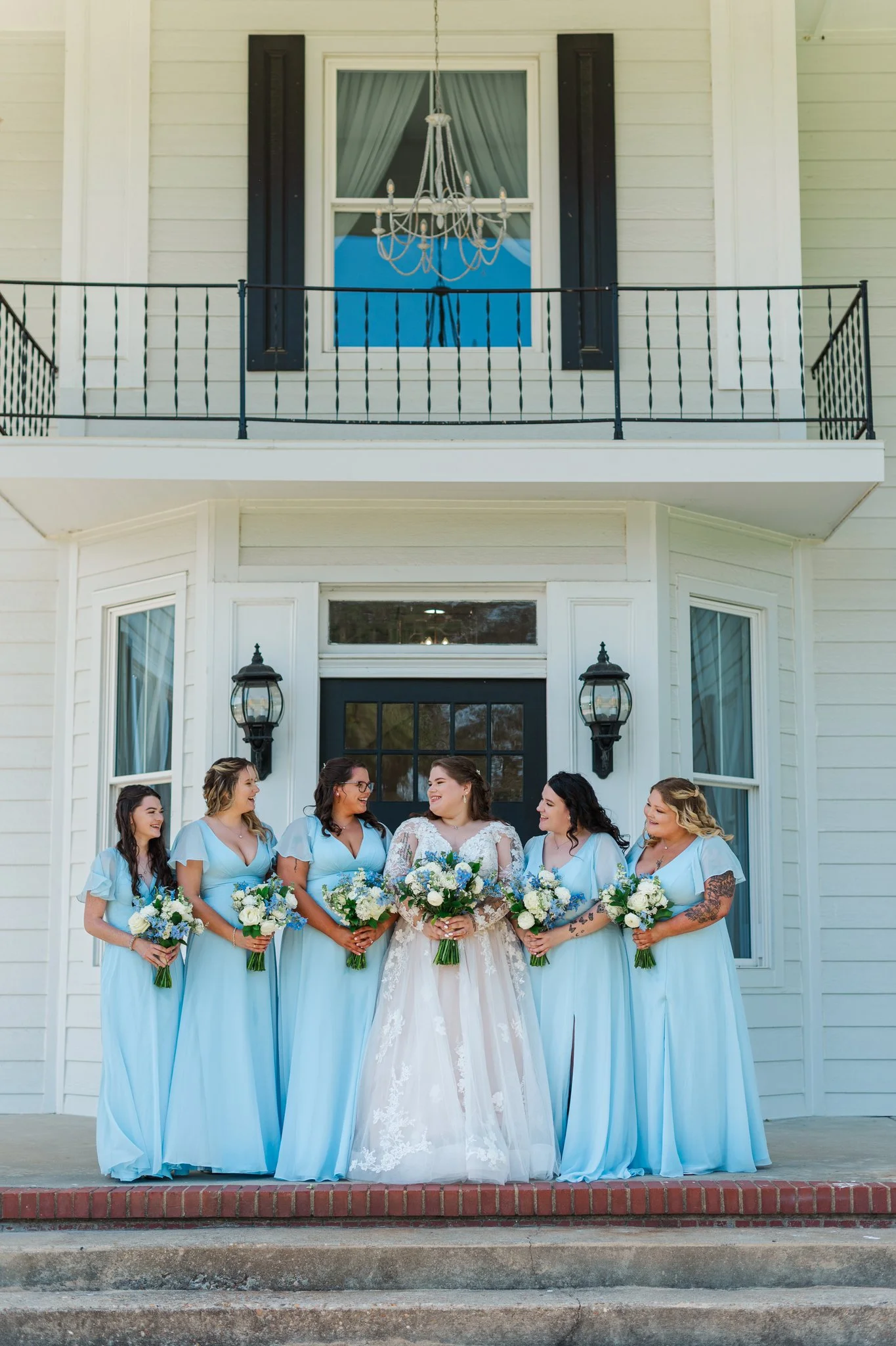 Bride and five bridesmaids standing on front steps of a white house, each holding a bouquet of white and blue flowers, with the bride in a white lace gown and the bridesmaids in blue dresses.