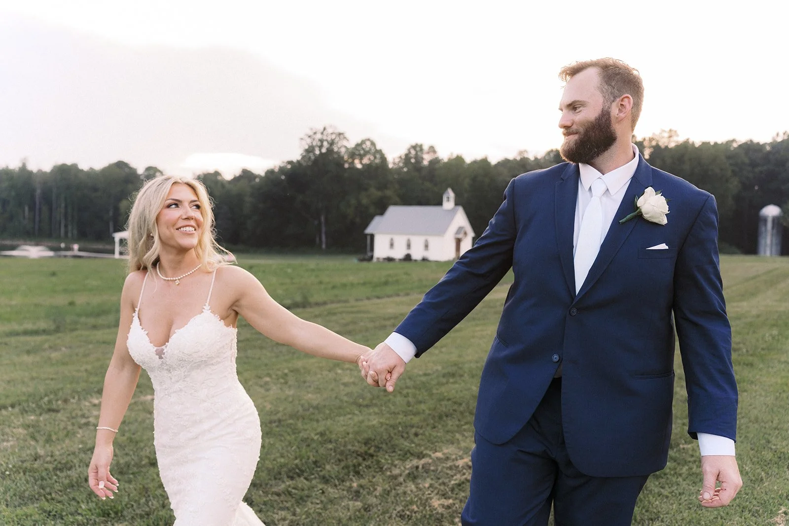 A newly married couple holding hands outdoors on a grassy field with a small white chapel and trees in the background. The bride is wearing a white lace wedding dress, and the groom is in a navy suit with a white shirt and boutonniere. The bride is s