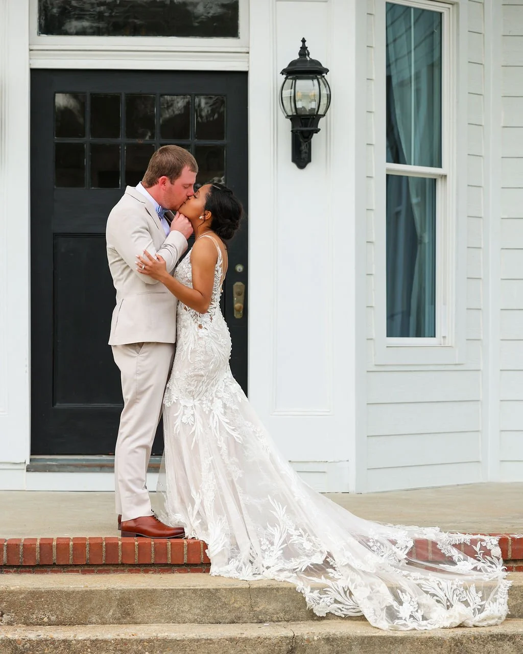 A newlywed couple sharing a kiss outside a house, with the bride in a white lace wedding gown and the groom in a light-colored suit, standing on a small brick step.