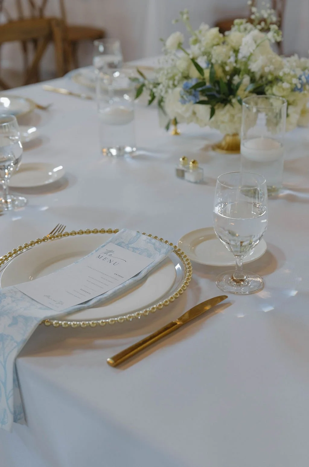 Elegant wedding reception table setup with white linens, gold cutlery, a white plate with beaded gold rim, a menu card, a napkin, flower centerpiece with white and light blue flowers, and glasses of water.