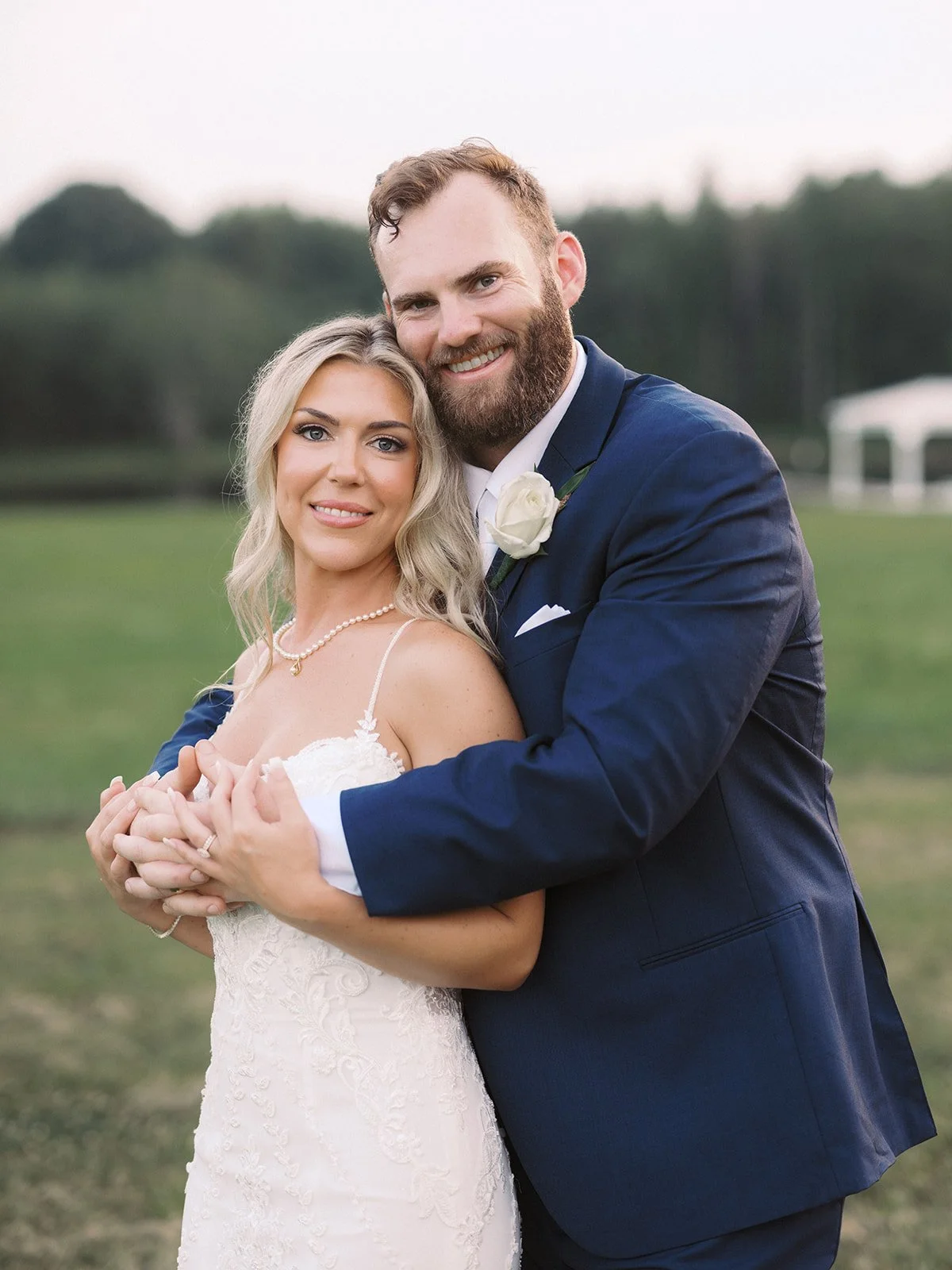 A bride and groom embrace outdoors on their wedding day, smiling at the camera. The bride is wearing a white lace wedding gown with spaghetti straps and a pearl necklace. The groom is dressed in a navy blue suit with a white shirt and a white boutonn