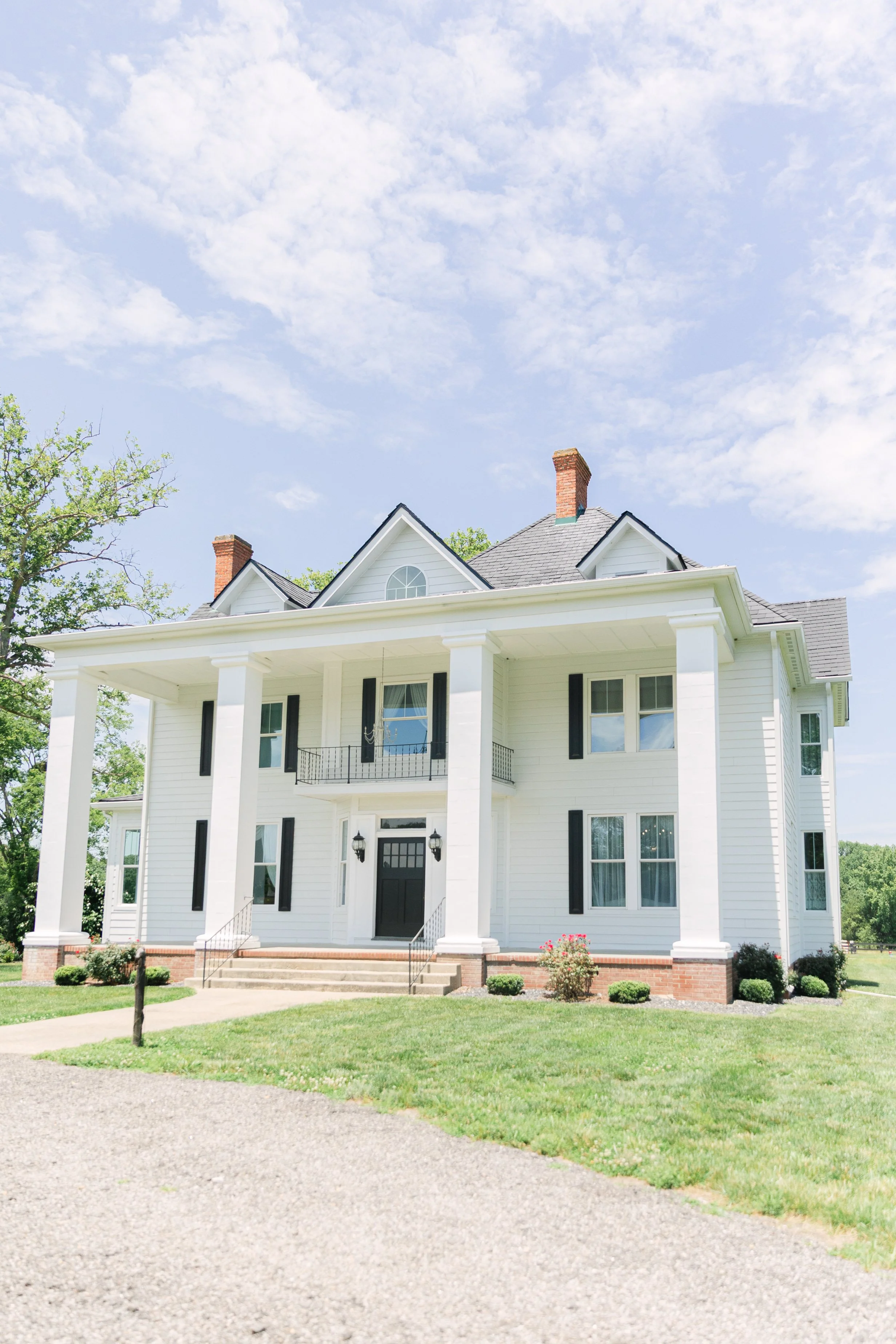 A large white two-story house with black shutters, a front porch, and a green lawn, under a partly cloudy sky.