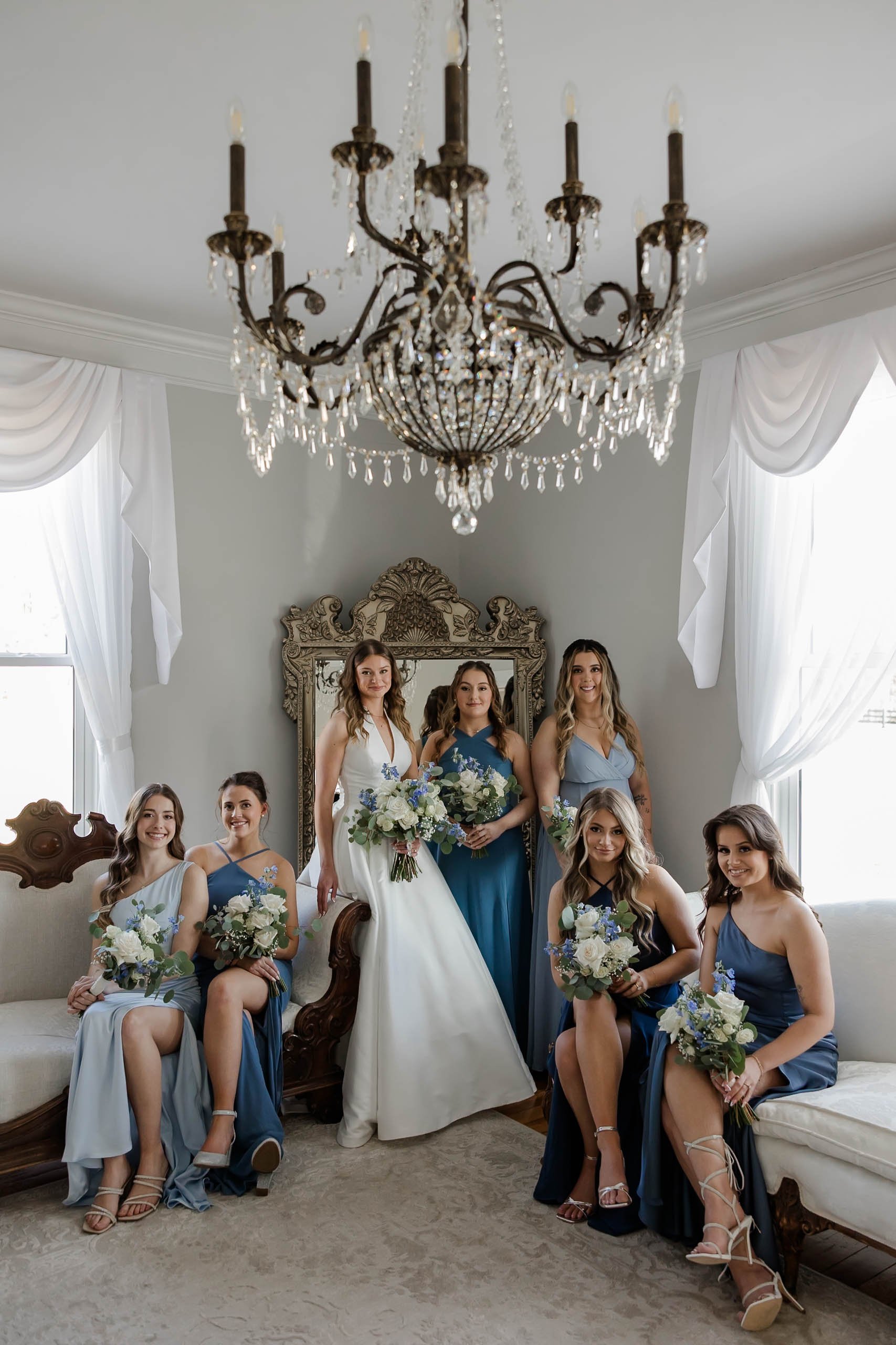 A bride and six bridesmaids posing in a vintage living room with large windows, white curtains, a chandelier, and an ornate mirror. The bride is in a white gown, and the bridesmaids are in blue dresses, holding bouquets of white and blue flowers.