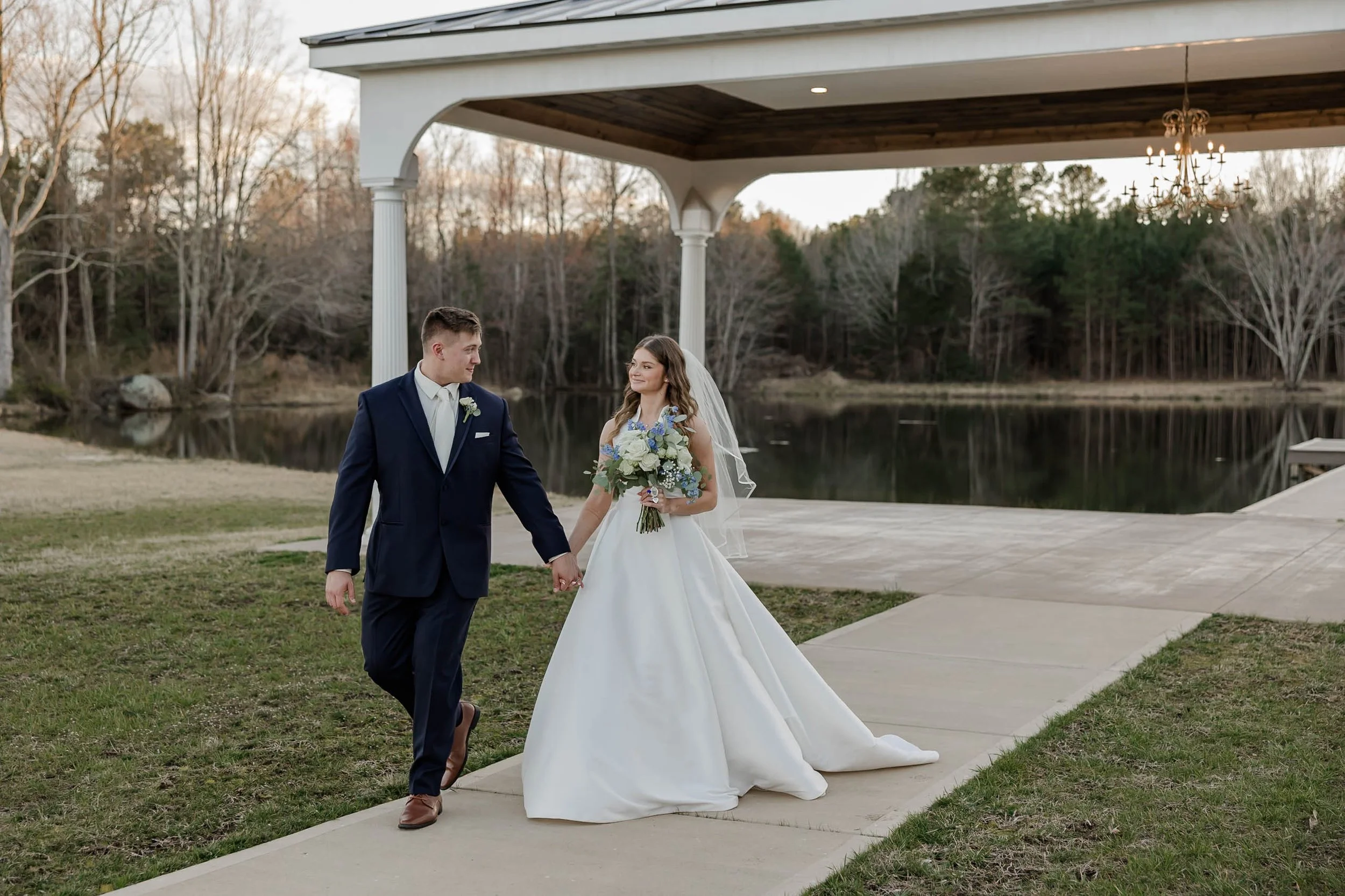 A bride and groom holding hands and walking outdoors near a lake, with trees and a covered porch with a chandelier in the background.