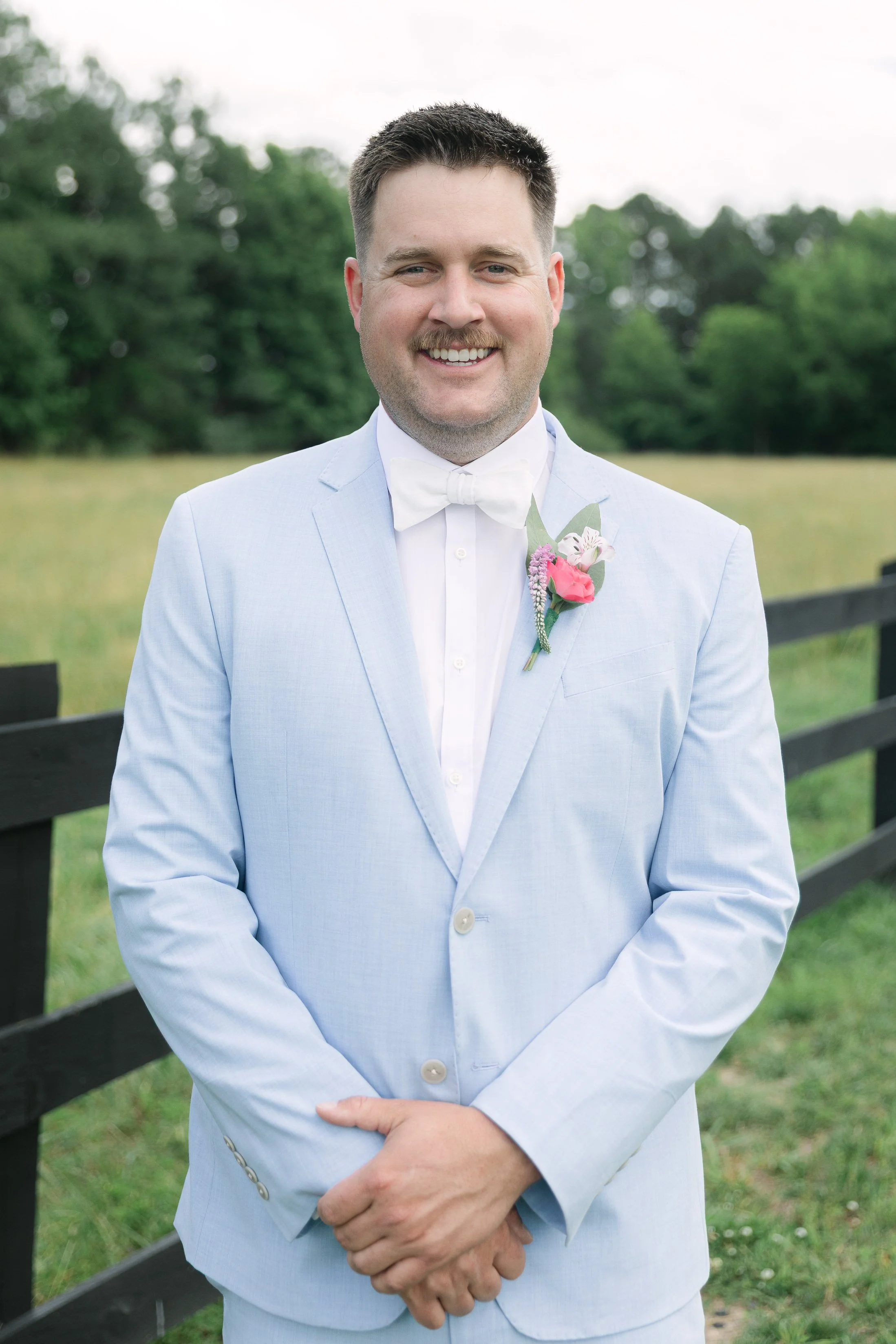A man dressed in a light blue suit with a white shirt and bow tie, standing outdoors near a black fence, smiling with a pink boutonniere on his jacket lapel, with green trees and grass in the background.