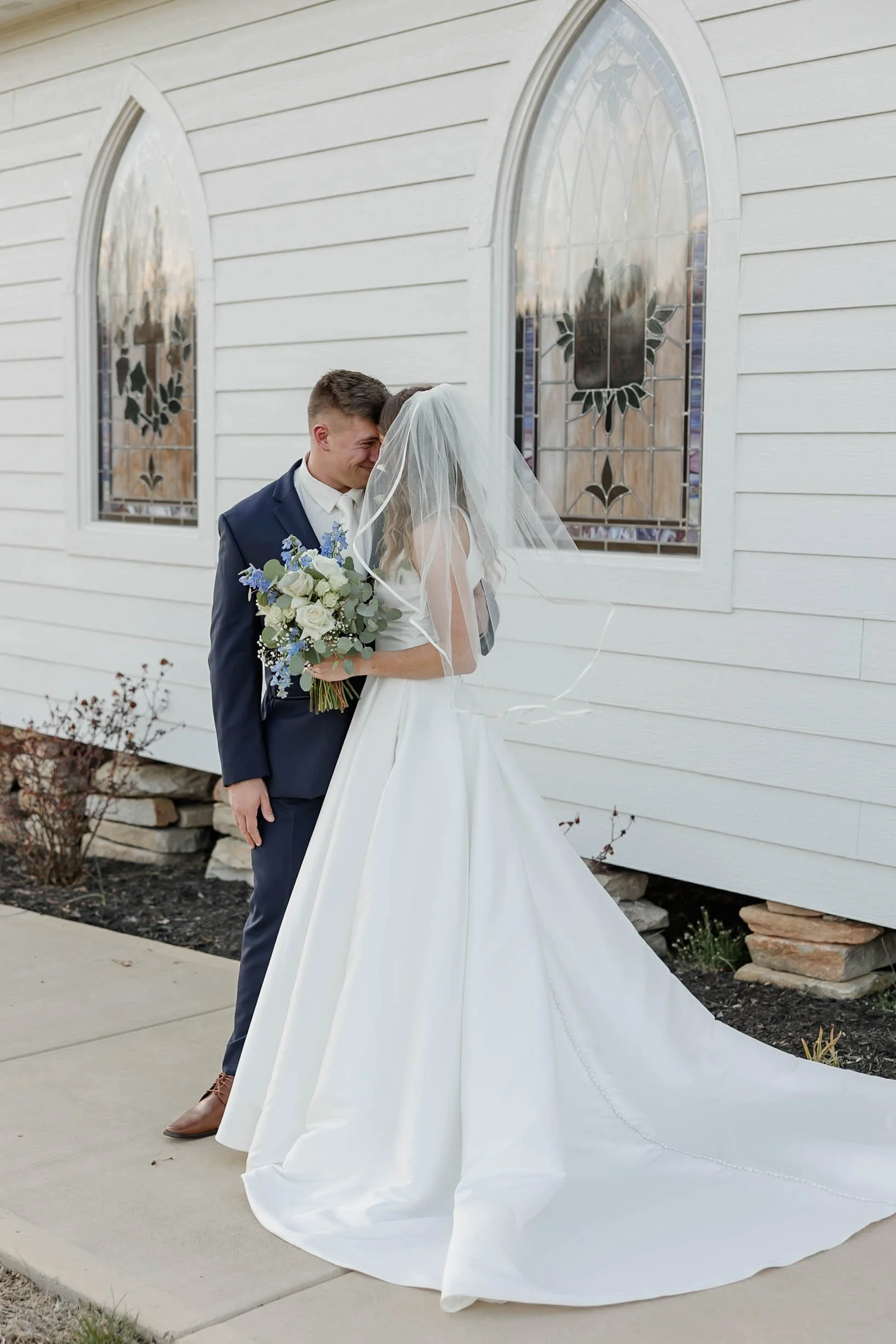 A bride and groom in wedding attire sharing a tender moment in front of a white building with stained glass windows.