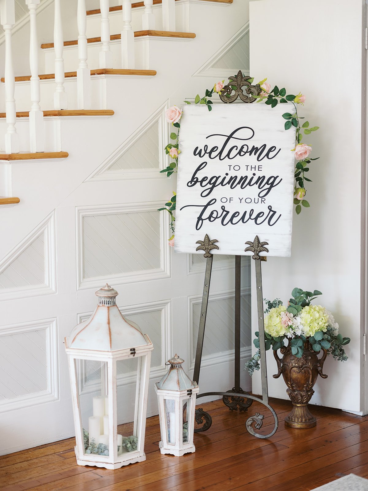 Decorative welcome sign on an easel with pink roses, a large vase of white and pale pink flowers, and two white lanterns with candles on a wooden floor near a staircase.