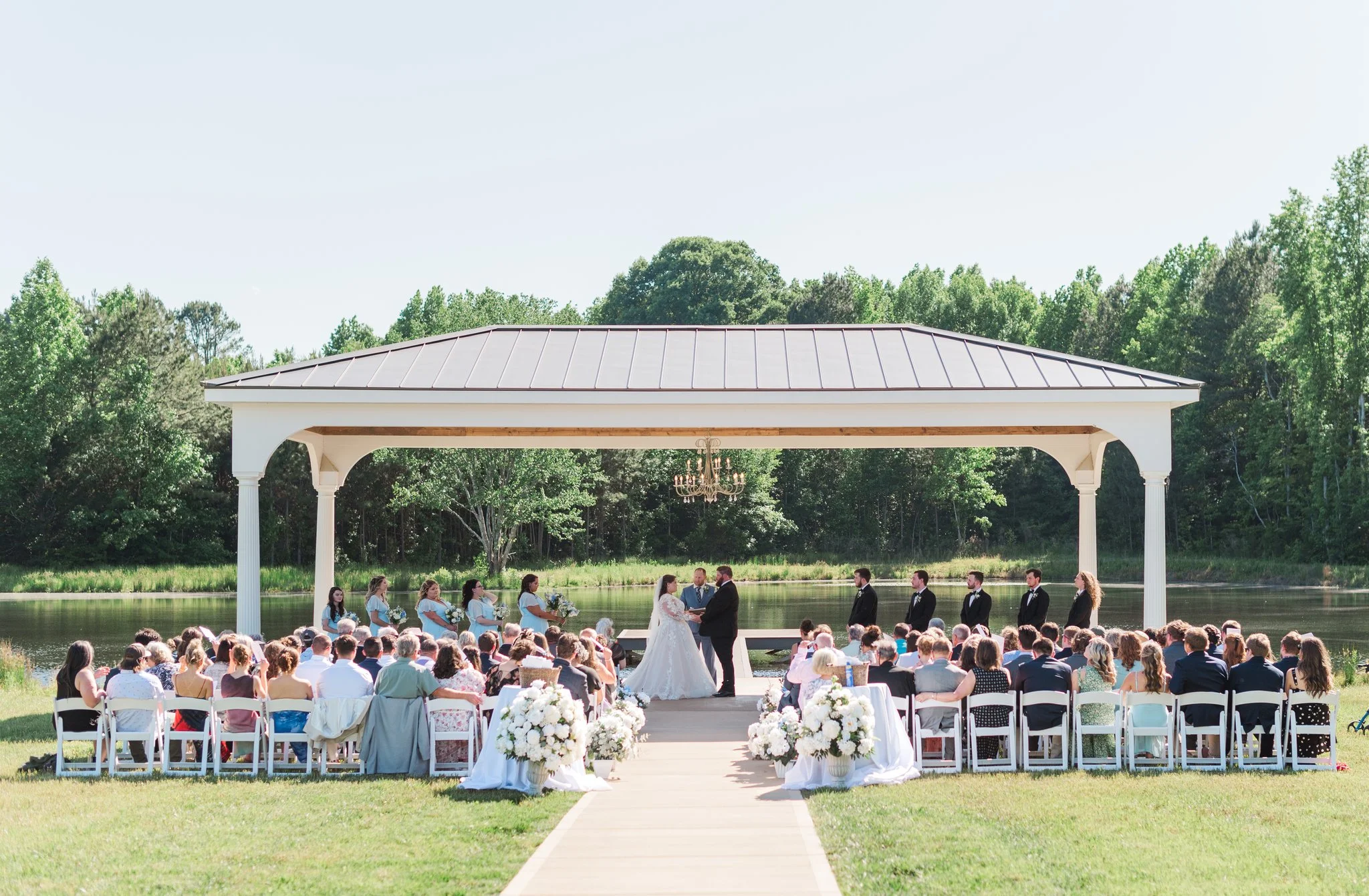 Outdoor wedding ceremony on a pond, with a white canopy, bride and groom exchanging vows, surrounded by seated guests and bridal party, during daytime.