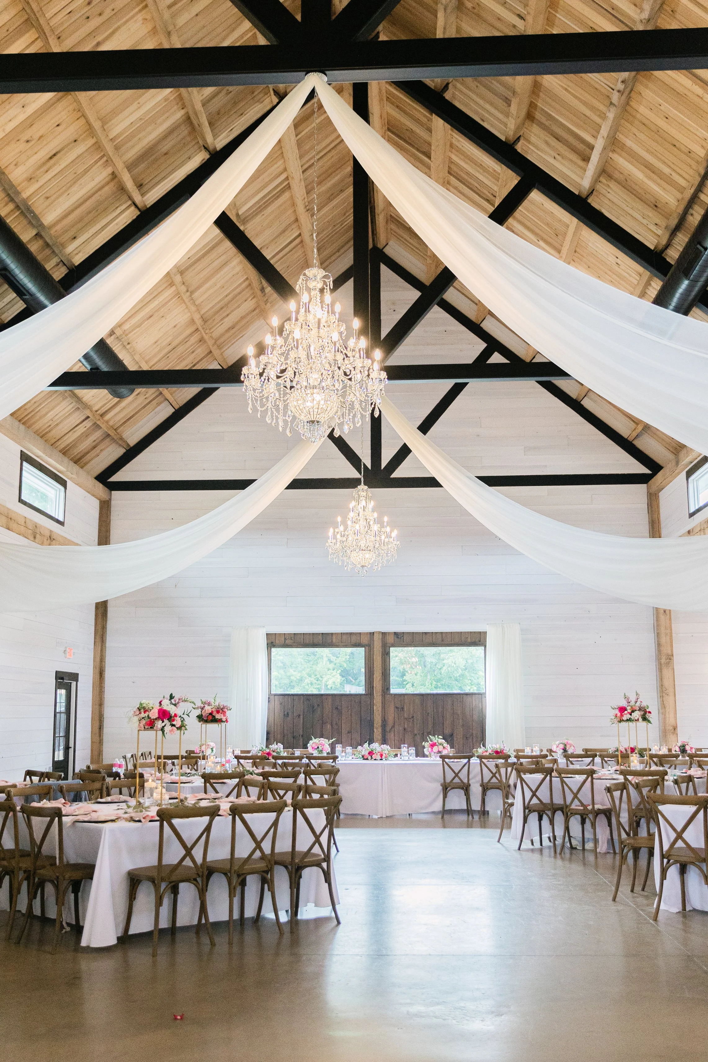 Wedding reception hall decorated with white drapery, chandeliers, and pink floral centerpieces on round tables with wooden chairs.