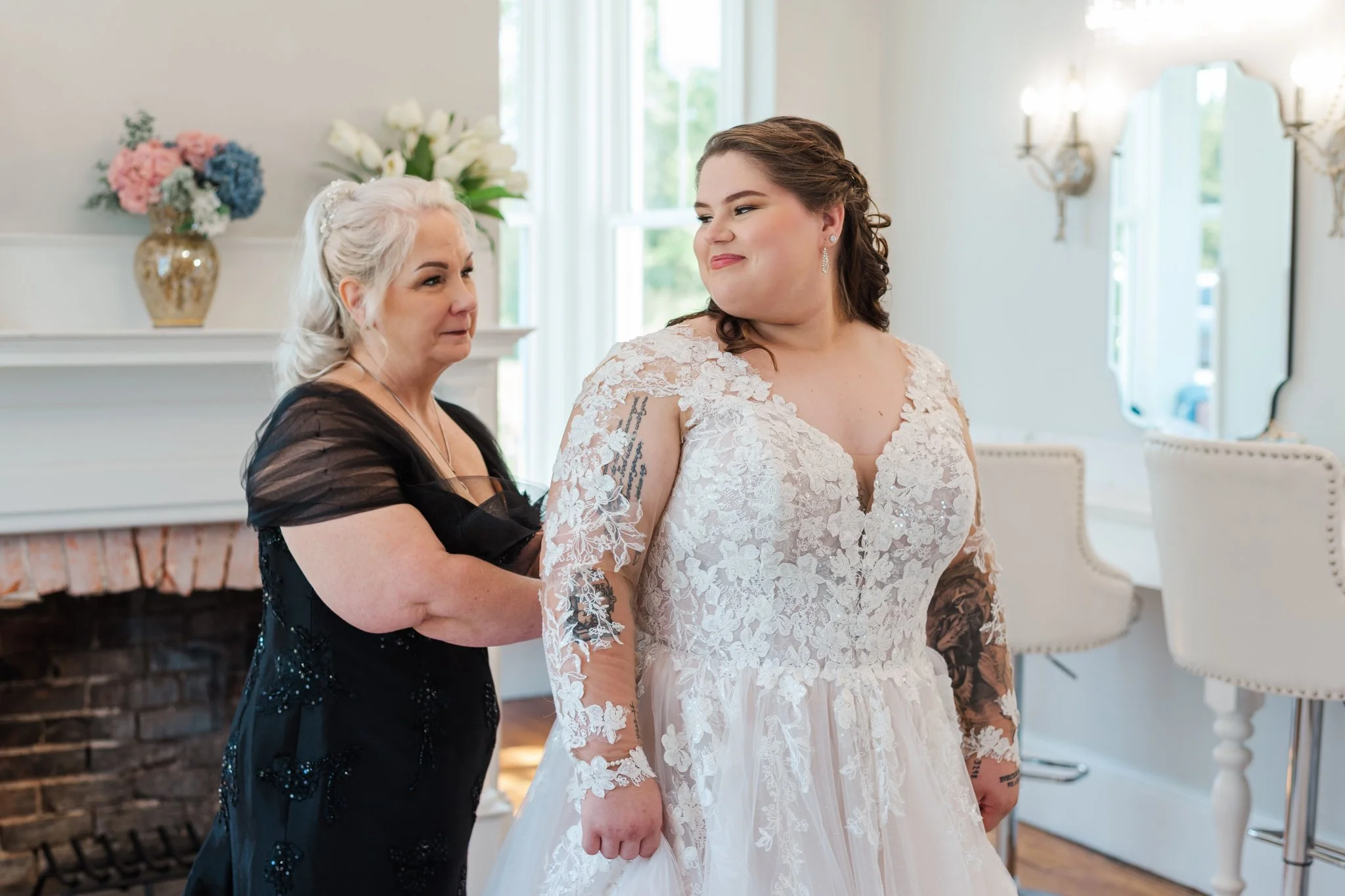 A bride in a wedding dress standing in front of an older woman, who appears to be adjusting or helping her with the dress. The room has white walls, a fireplace with floral decorations on the mantel, and a window with natural light.