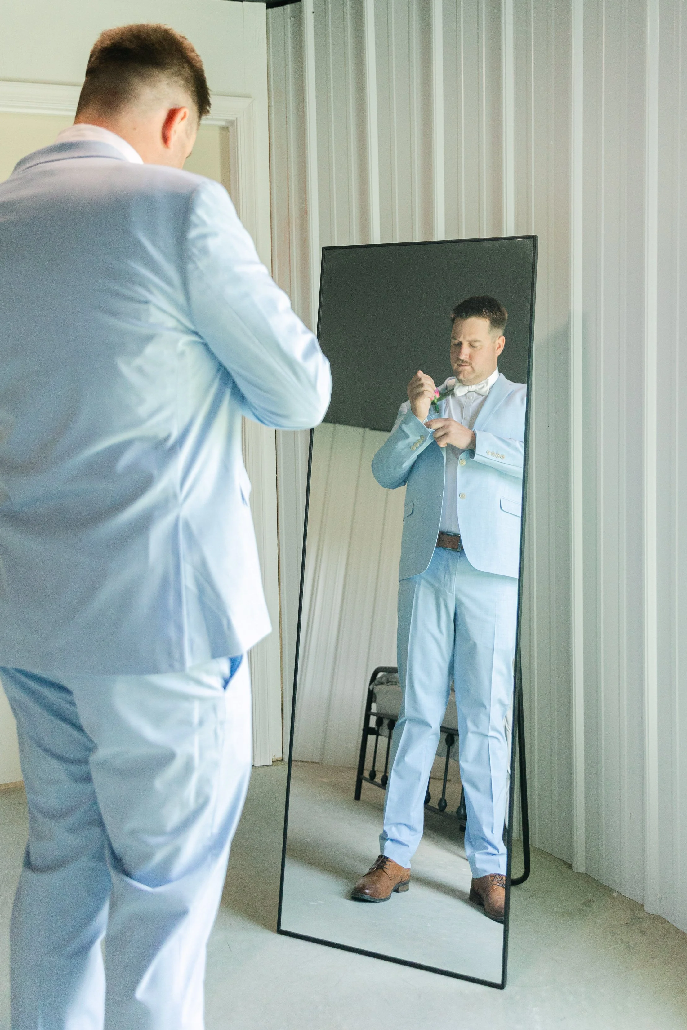 A man in a light blue suit is standing in front of a full-length mirror, adjusting his bow tie. The man is reflected in the mirror, showing him from the front. The room has white-paneled walls and a beige carpet.