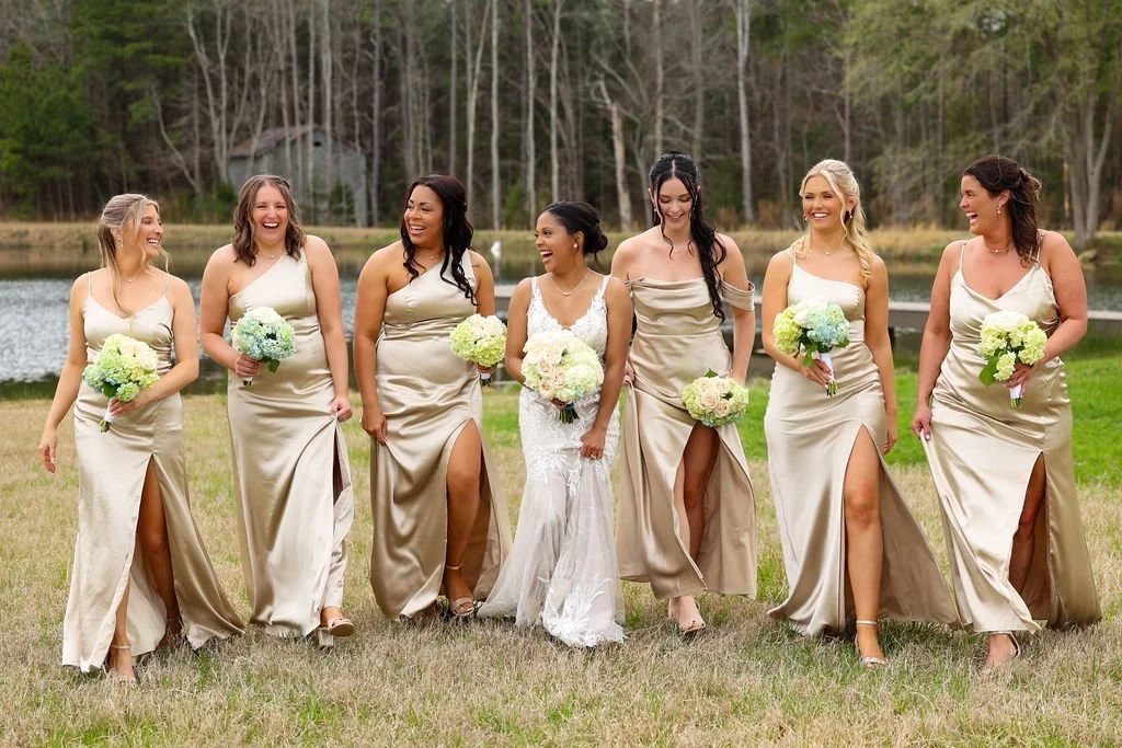 A bride and seven bridesmaids walking outdoors near a lake, holding bouquets of flowers, dressed in matching cream-colored dresses.