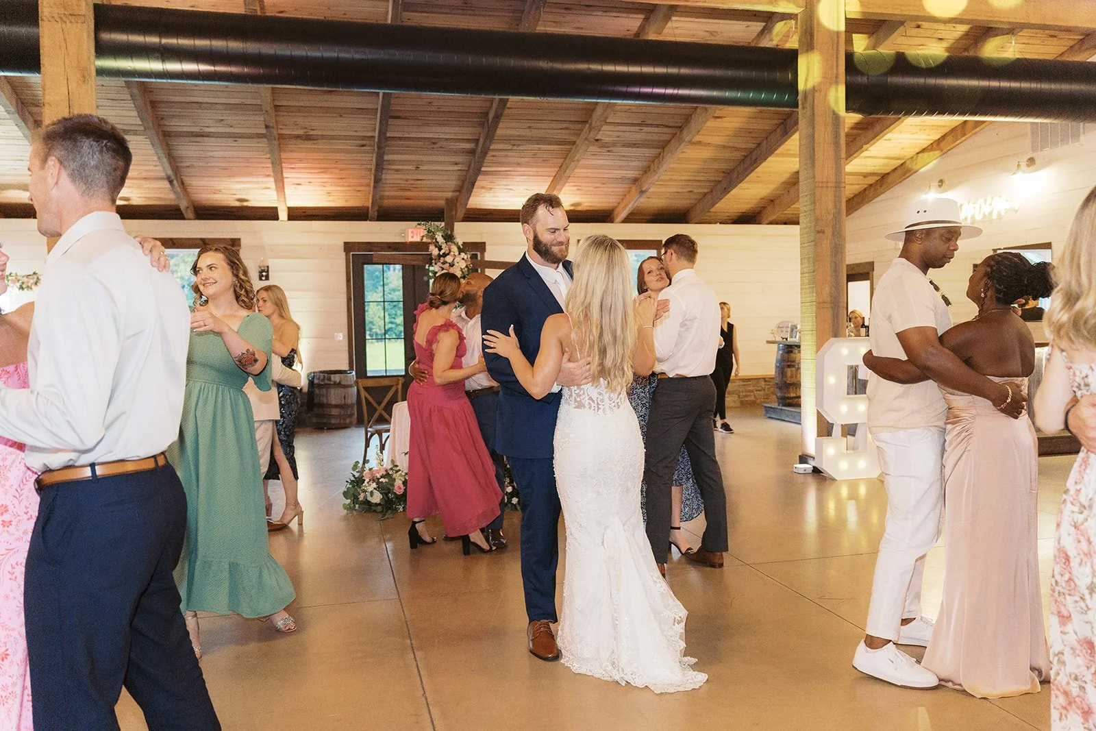 Couples dancing at a wedding reception in a rustic indoor venue with wooden beams and decor.