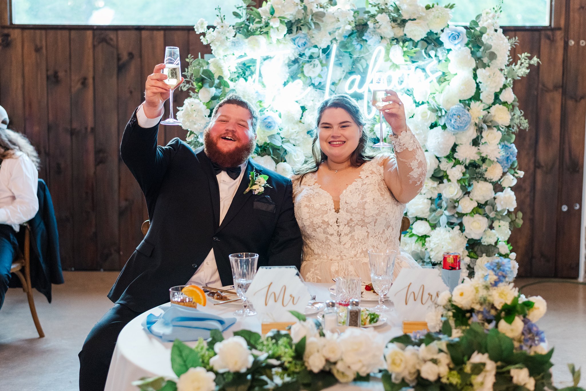 A newlywed couple, a man in a tuxedo and a woman in a lace wedding dress, sitting at a wedding reception table with floral decorations and toasts, each holding a glass of champagne and smiling.