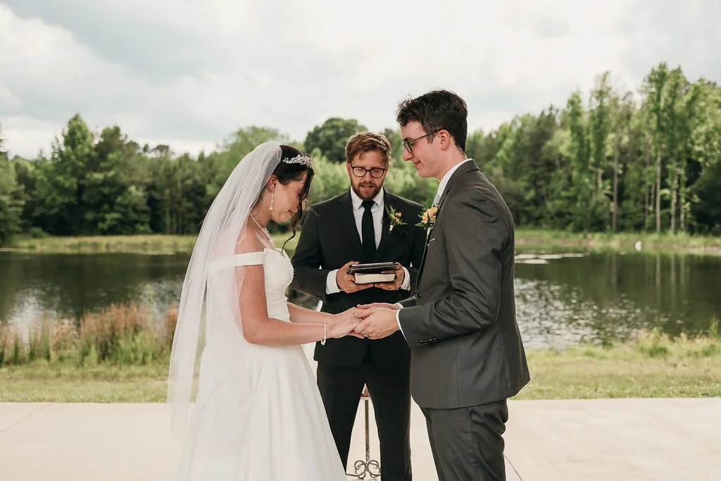A bride and groom holding hands during an outdoor wedding ceremony near a lake, with officiant standing behind them holding a book, and trees in the background.