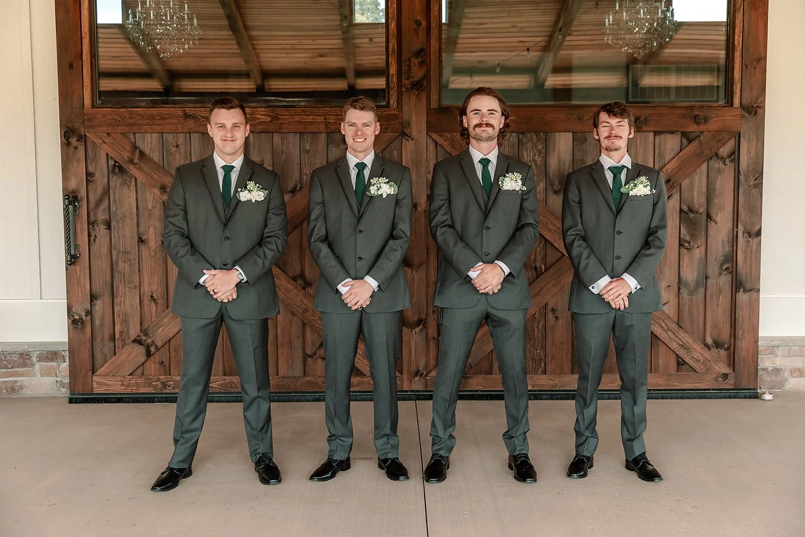 Four men in matching gray suits with white shirts, green ties, and boutonnières stand in a row in front of a wooden barn door with glass windows and chandeliers reflected in the glass.