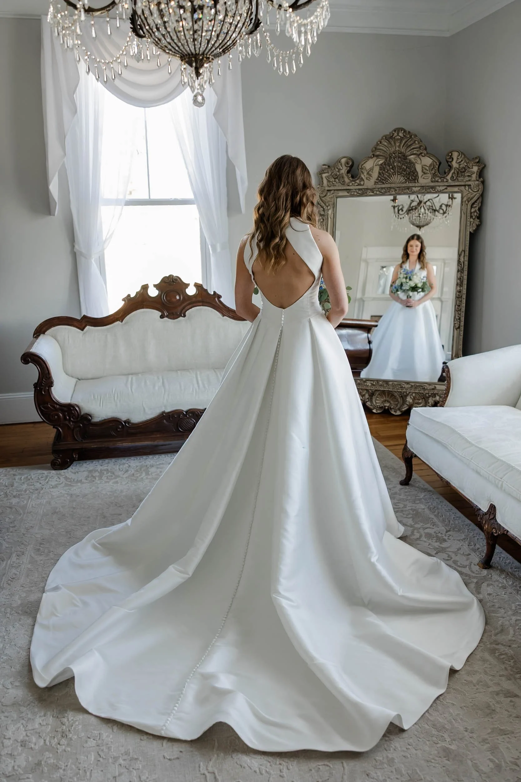 A bride in a white wedding dress standing in front of a large ornate mirror, holding a bouquet of flowers, in a decorated room with a chandelier and vintage furniture.