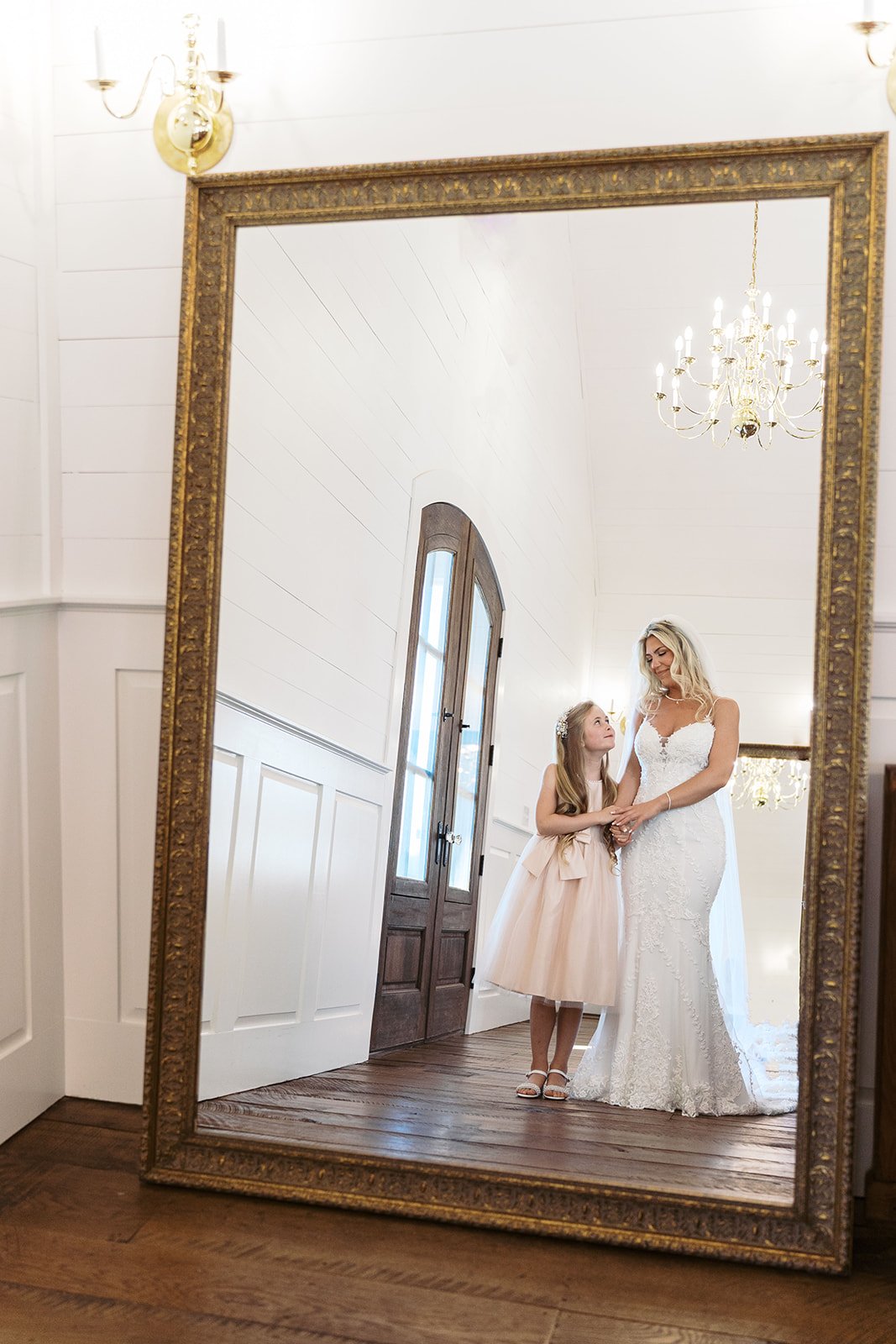 A bride and a young girl are reflected in a large ornate mirror inside a bright, elegant room with white walls, wood floors, a chandelier, and a chandelier-shaped wall sconce.