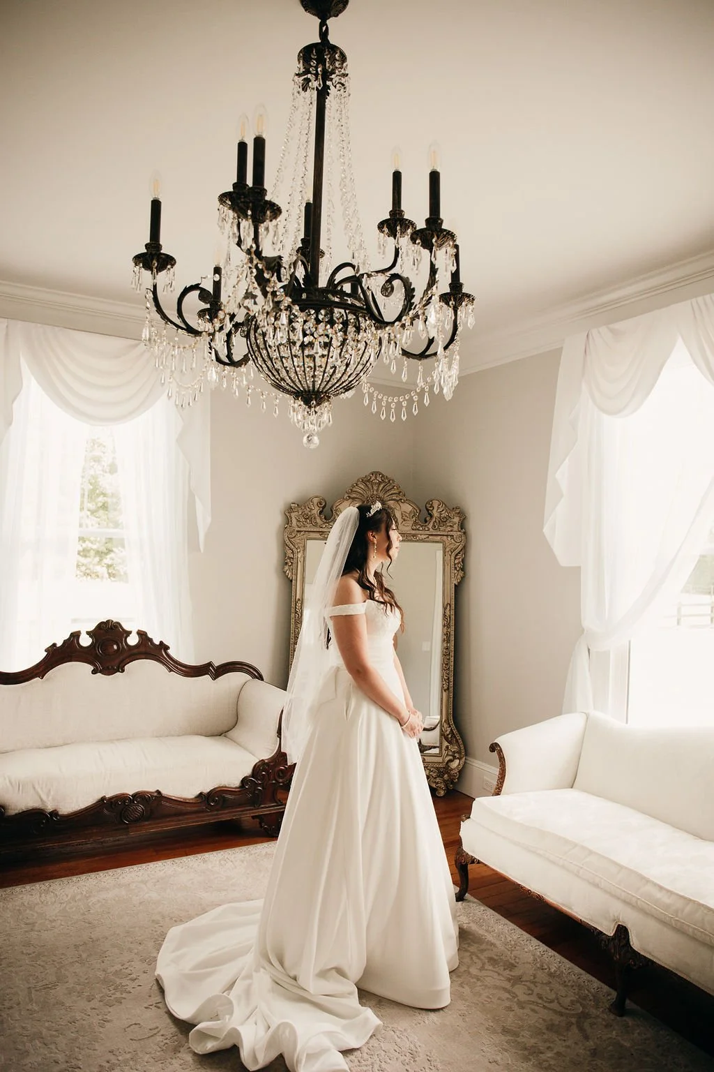 Bride in a white wedding dress standing in a bright, elegant room with chandeliers, vintage furniture, and white curtains.