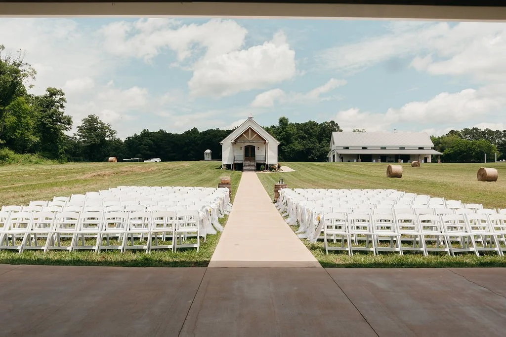 An outdoor wedding setup with white folding chairs arranged on either side of a wooden aisle leading to a small white church in a grassy field with hay bales and a barn in the background, under a partly cloudy sky.