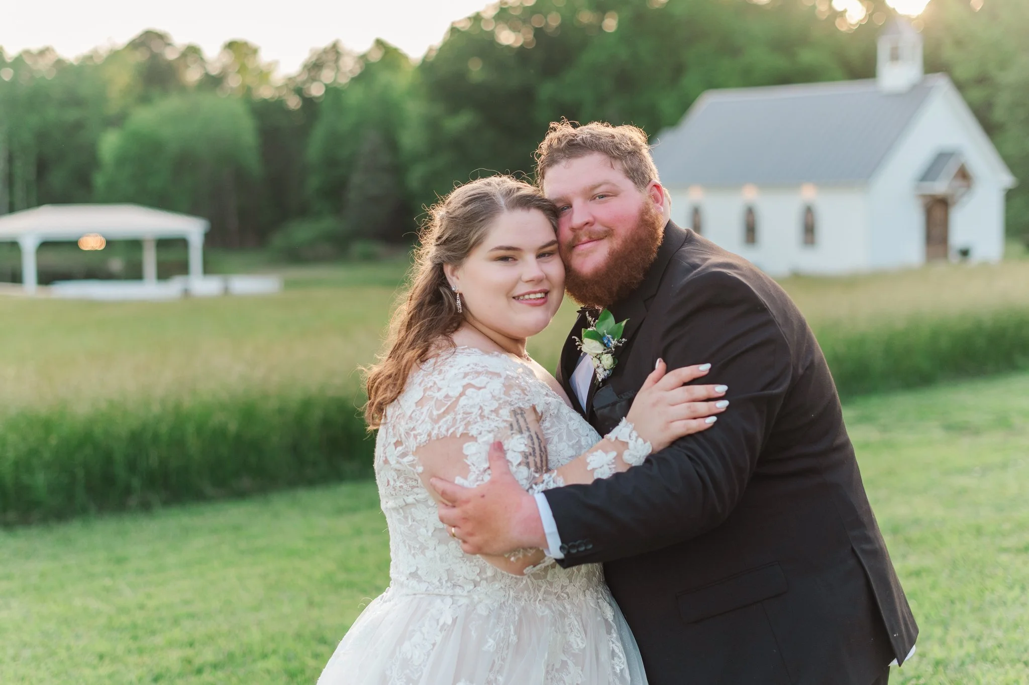 A bride and groom embracing outdoors on their wedding day with a white church and a gazebo in the background.