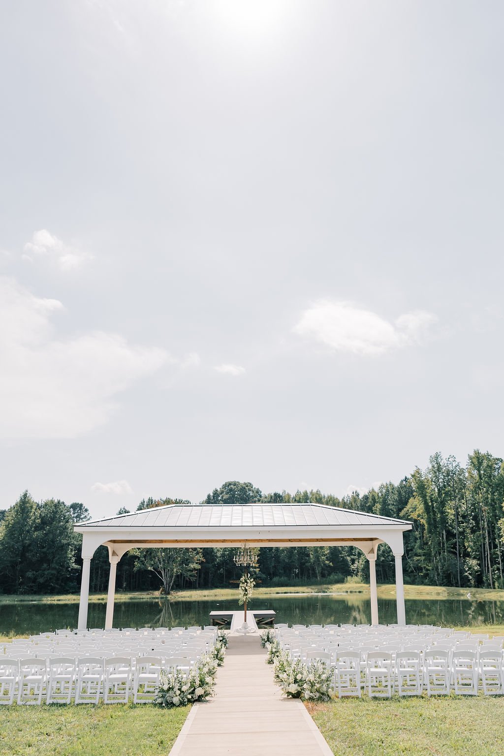 Outdoor wedding ceremony setup with white chairs on either side of a wooden aisle leading to a decorated arch near a pond, with greenery and trees in the background under a partly cloudy sky.