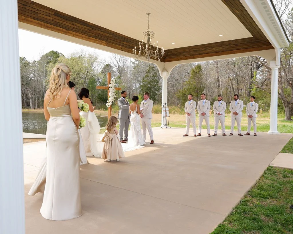 A wedding ceremony taking place outdoor beneath a covered patio near a lake, with the bride and groom standing at the altar, surrounded by bridesmaids and groomsmen in light-colored suits, and a young girl holding flowers.