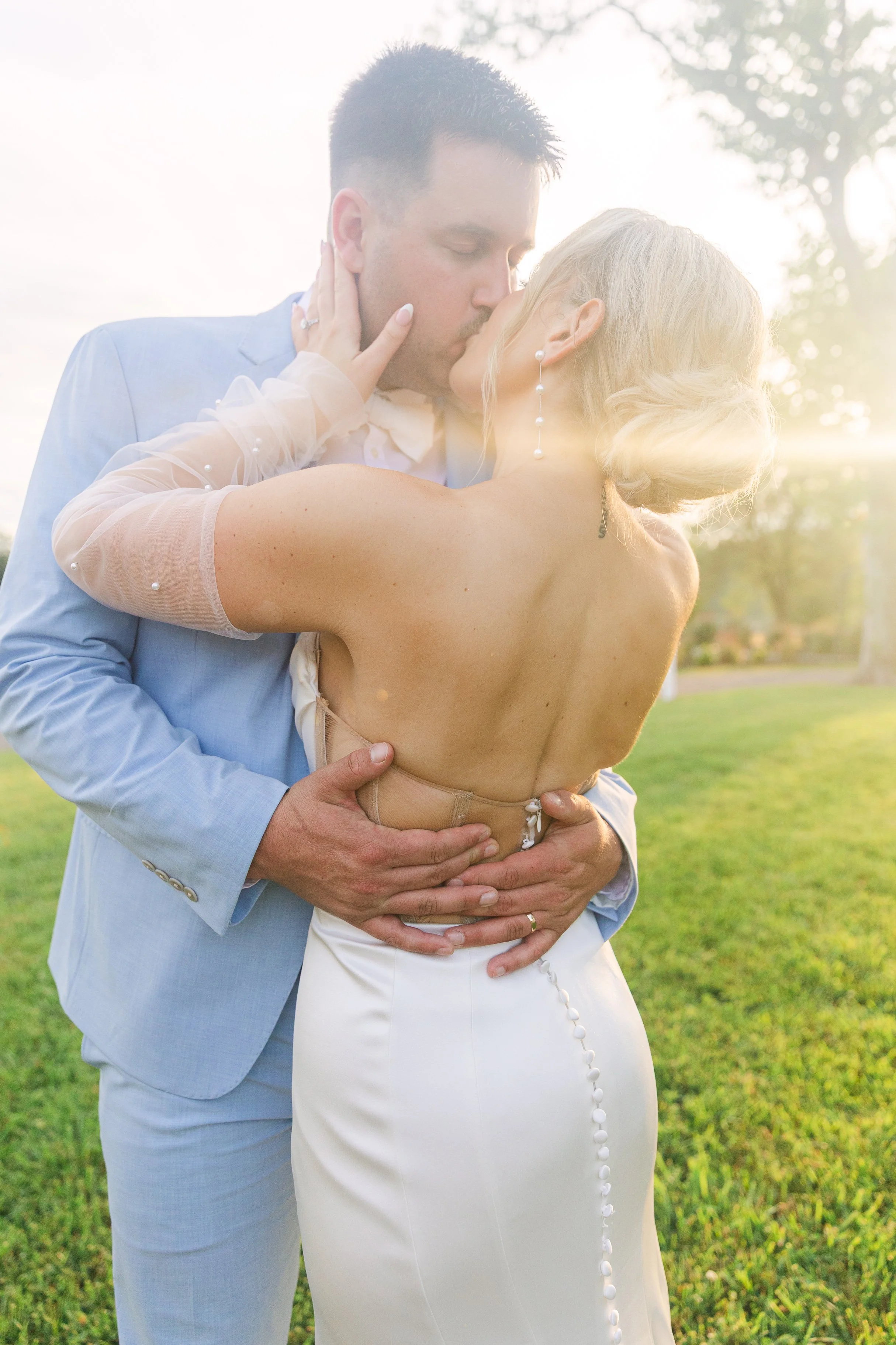 A newlywed couple sharing a kiss outdoors in sunlight. The groom is wearing a light blue suit, and the bride is wearing a backless wedding dress with buttons down the back and pearl accents. They are embracing on a grassy area with trees in the backg