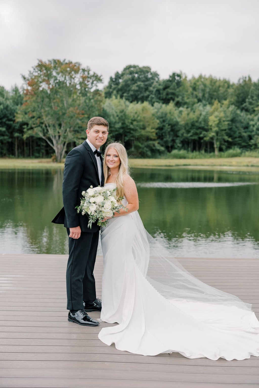 A newlywed couple stands on a wooden dock by a lake, smiling and holding a bouquet of flowers, with trees and water in the background.