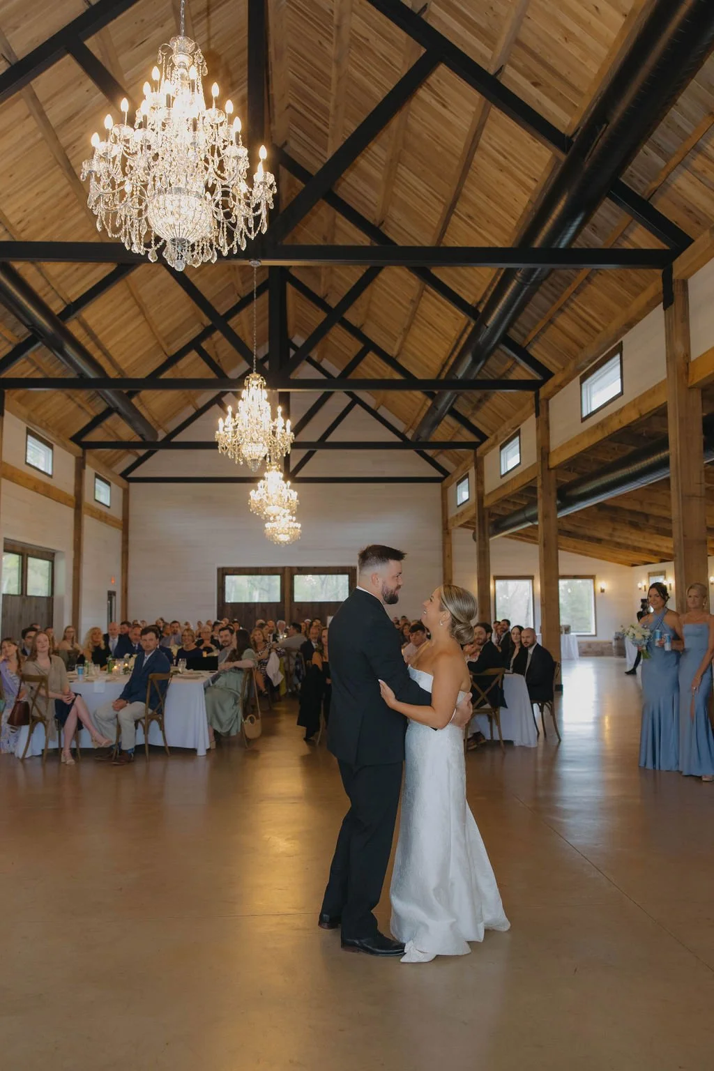 A bride and groom share their first dance at a wedding reception in a rustic venue with wooden beams and large chandeliers.